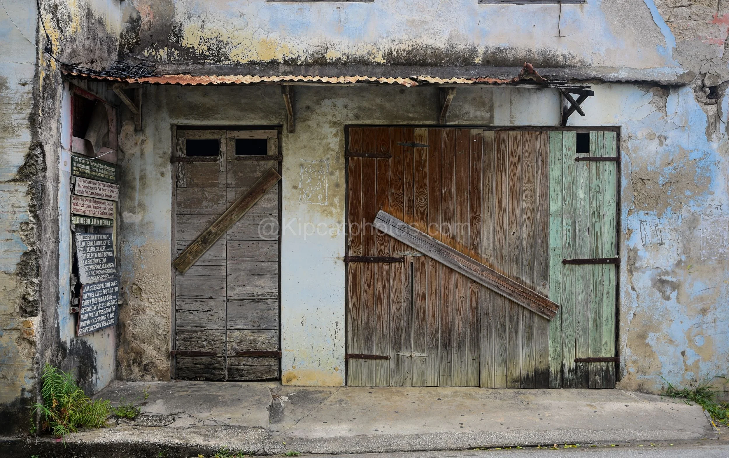 A weathered building facade in Speightstown, Barbados, with boarded wooden doors and hand-painted scripture signs. Kipcatphotos.com

A crumbling plastered facade in Speightstown shows decades of tropical weathering, with mismatched timber doors brace