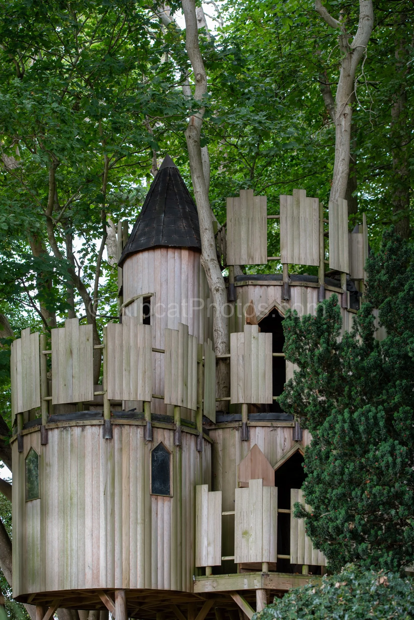 A large multi-turret castle-style timber treehouse sits high in the trees at Broughton Grange, Oxfordshire, its timber towers, battlements and pointed roof just visible through the green summer canopy. KipcatPhotos.com.

Hidden among the trees at Bro