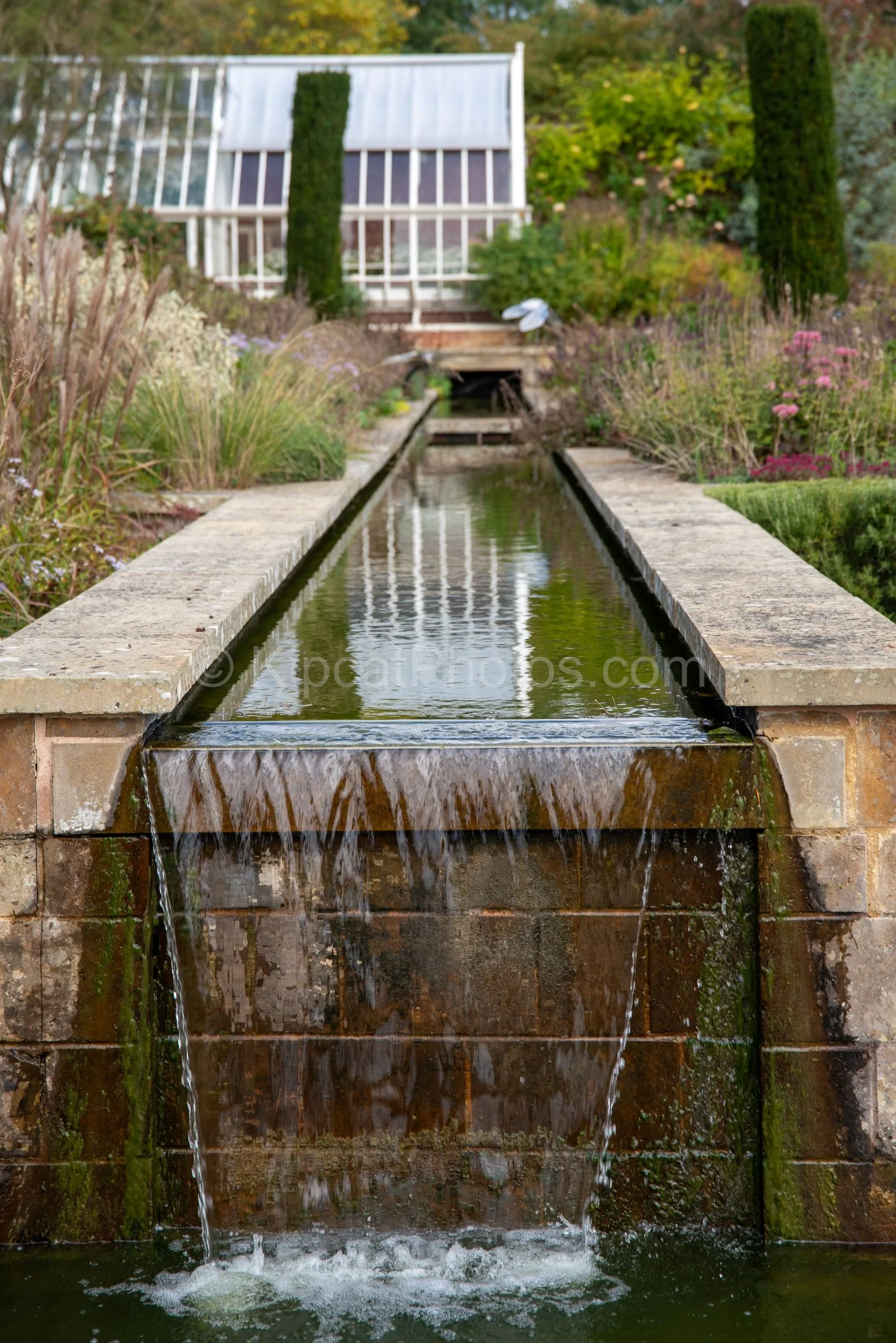 A long stone rill water feature overflows in a sheet waterfall cascade at Broughton Grange, Oxfordshire, with the Victorian glasshouse, columnar yew topiary and mixed perennial borders visible beyond. KipcatPhotos.com.

The stone rill at Broughton Gr