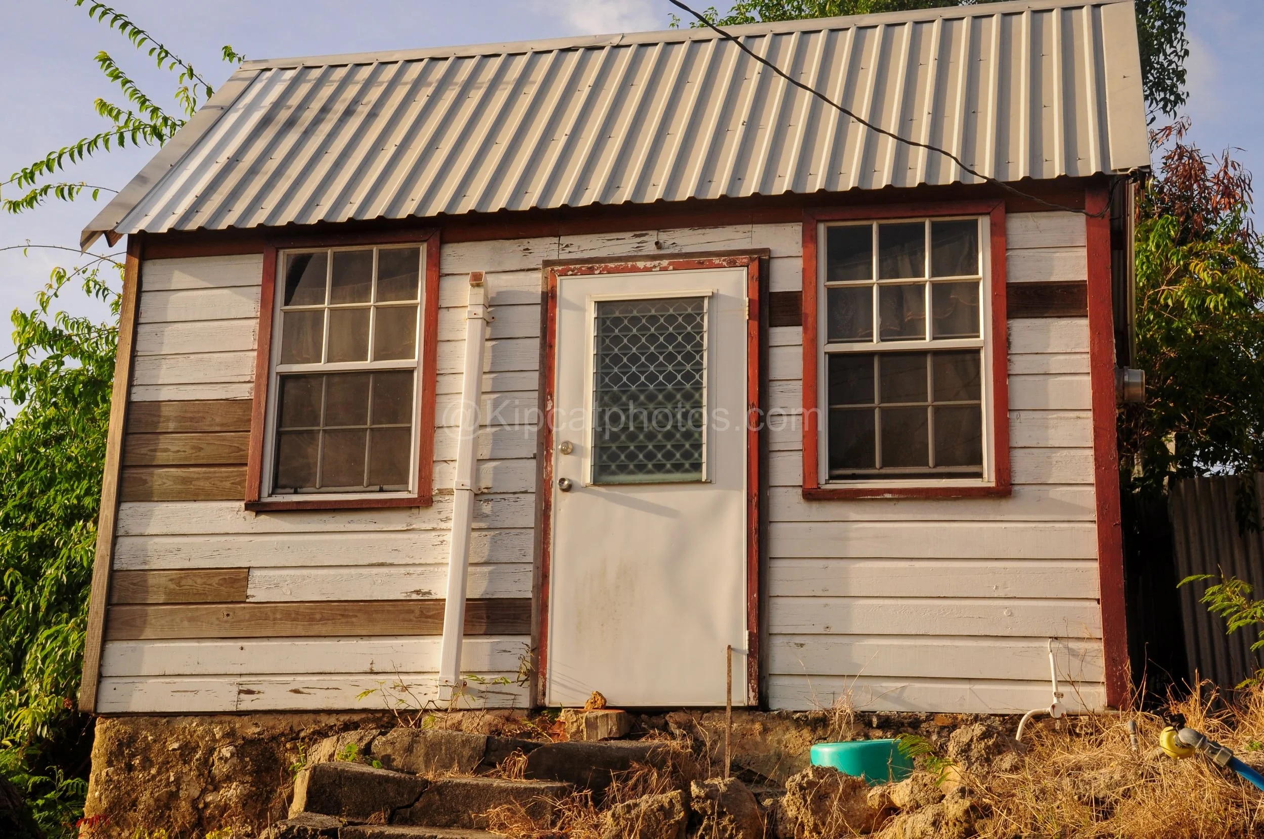 A weathered chattel house in Barbados with red-trimmed windows and mismatched replacement boards. Kipcatphotos.com

Patches of newer, darker timber stand out against this chattel house's sun-bleached white boards, its red-trimmed windows and door sho