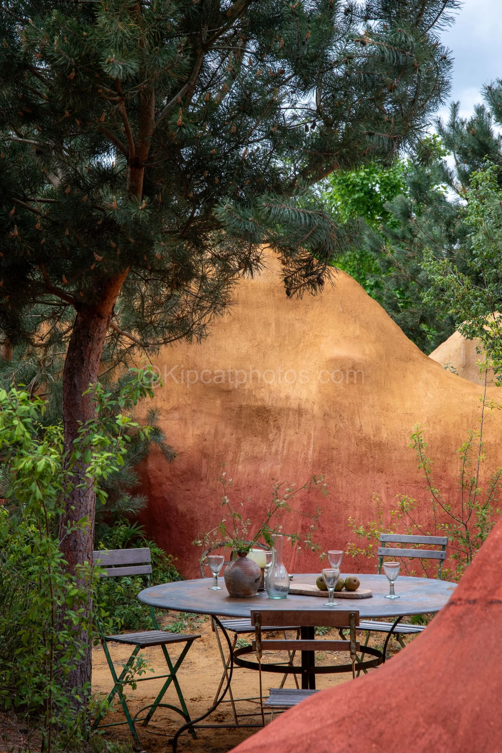 Outdoor dining table beneath pine trees against a red ochre wall in the James Basson garden at RHS Chelsea Flower Show 2026

A rustic round metal table set with glasses, a jug and a ceramic vase sits beneath tall pine trees, framed by a striking red 