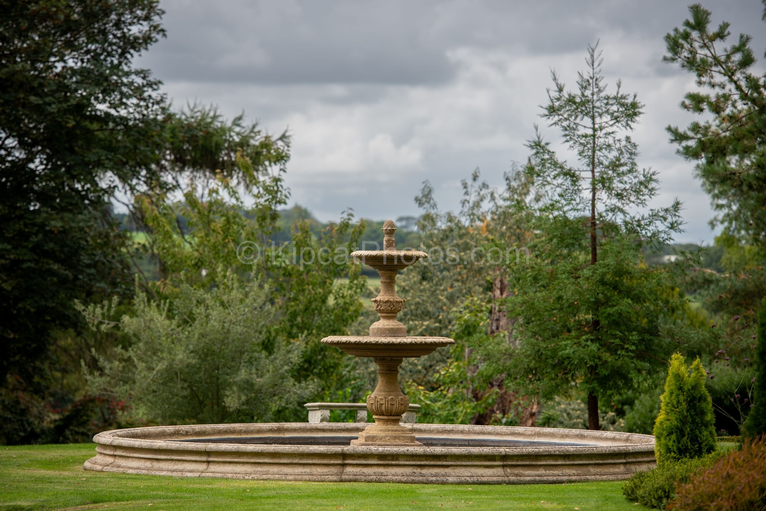 A tiered classical stone fountain stands on a formal lawn at Broughton Grange, Oxfordshire, with ornamental trees and the rolling Oxfordshire countryside visible beyond under a cloudy sky. KipcatPhotos.com.

The tiered classical fountain at Broughton