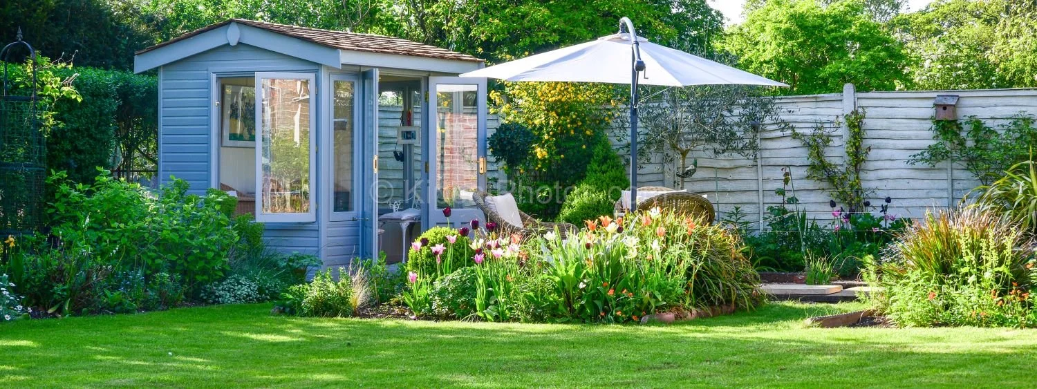 A sunny Berkshire garden in spring with a grey painted summerhouse, parasol and seating area, tulip and mixed flower borders on either side of a velvet lawn, and mature trees beyond. Kipcatphotos.com.

This cheerful spring panorama captures a well-lo