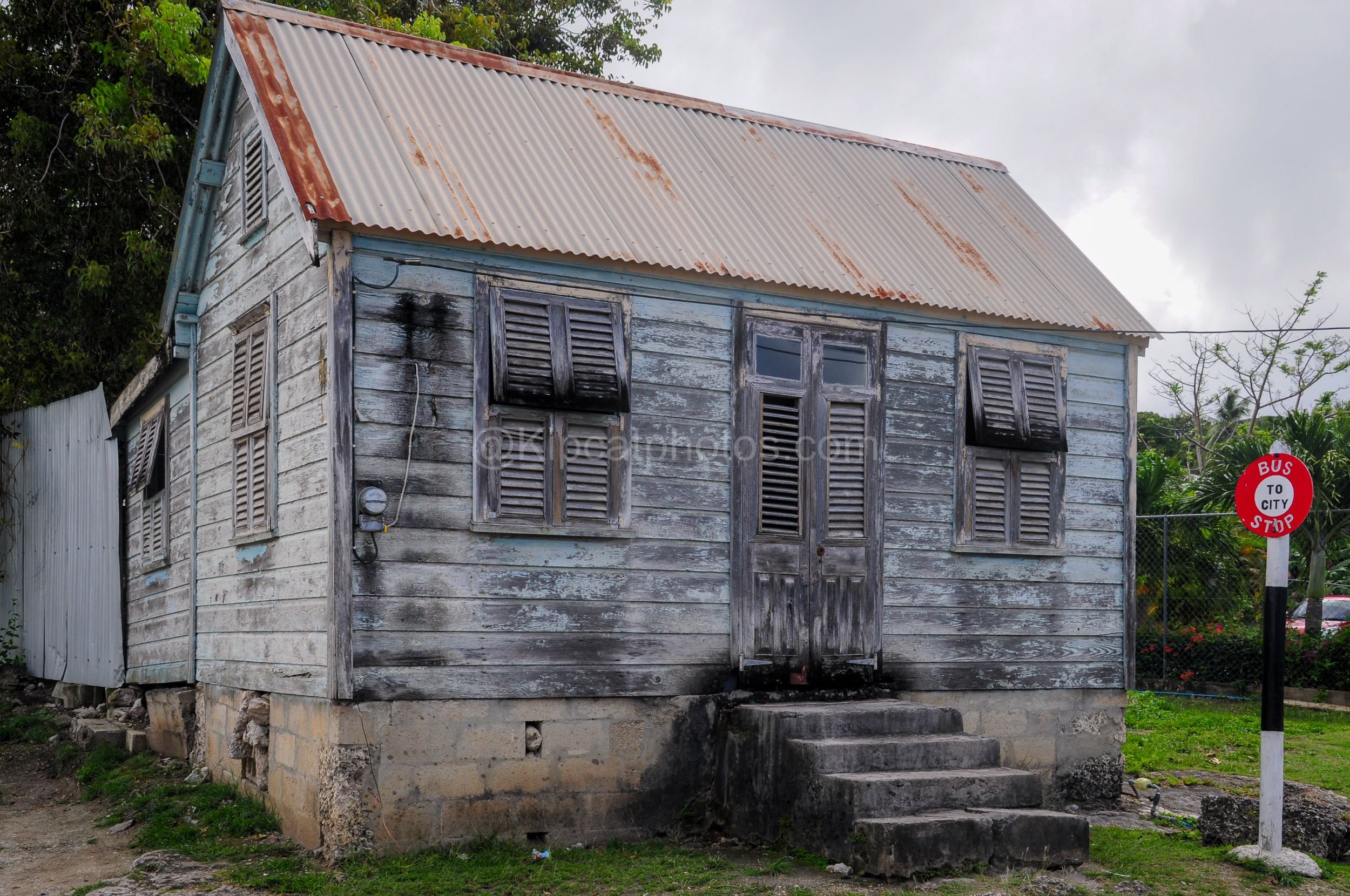 A pale blue chattel house in Barbados with weathered shutters, beside a "Bus to City" stop sign. Kipcatphotos.com

Streaked pale blue paint covers this chattel house's timber walls, its louvred shutters and panelled door showing years of exposure, wi