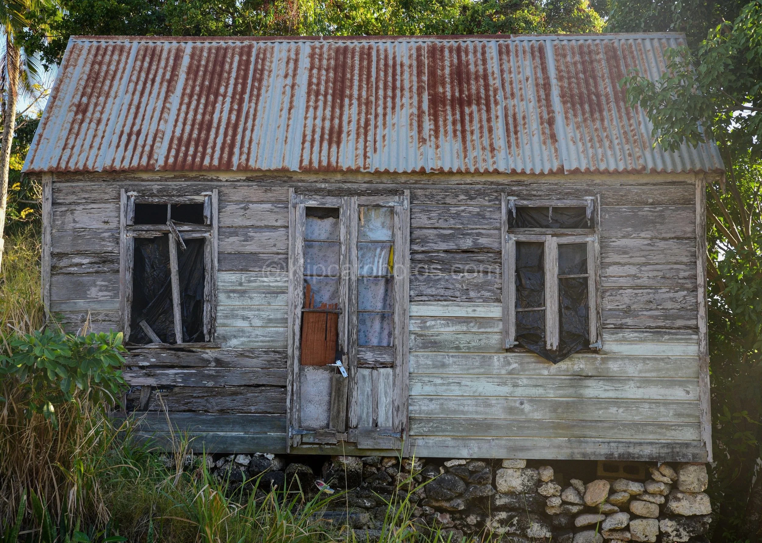 A heavily weathered chattel house in Barbados with broken windows covered in plastic sheeting. Kipcatphotos.com

Grey, sun-bleached boards and a broken window patched with plastic sheeting show this chattel house's advanced decay, its tattered lace c