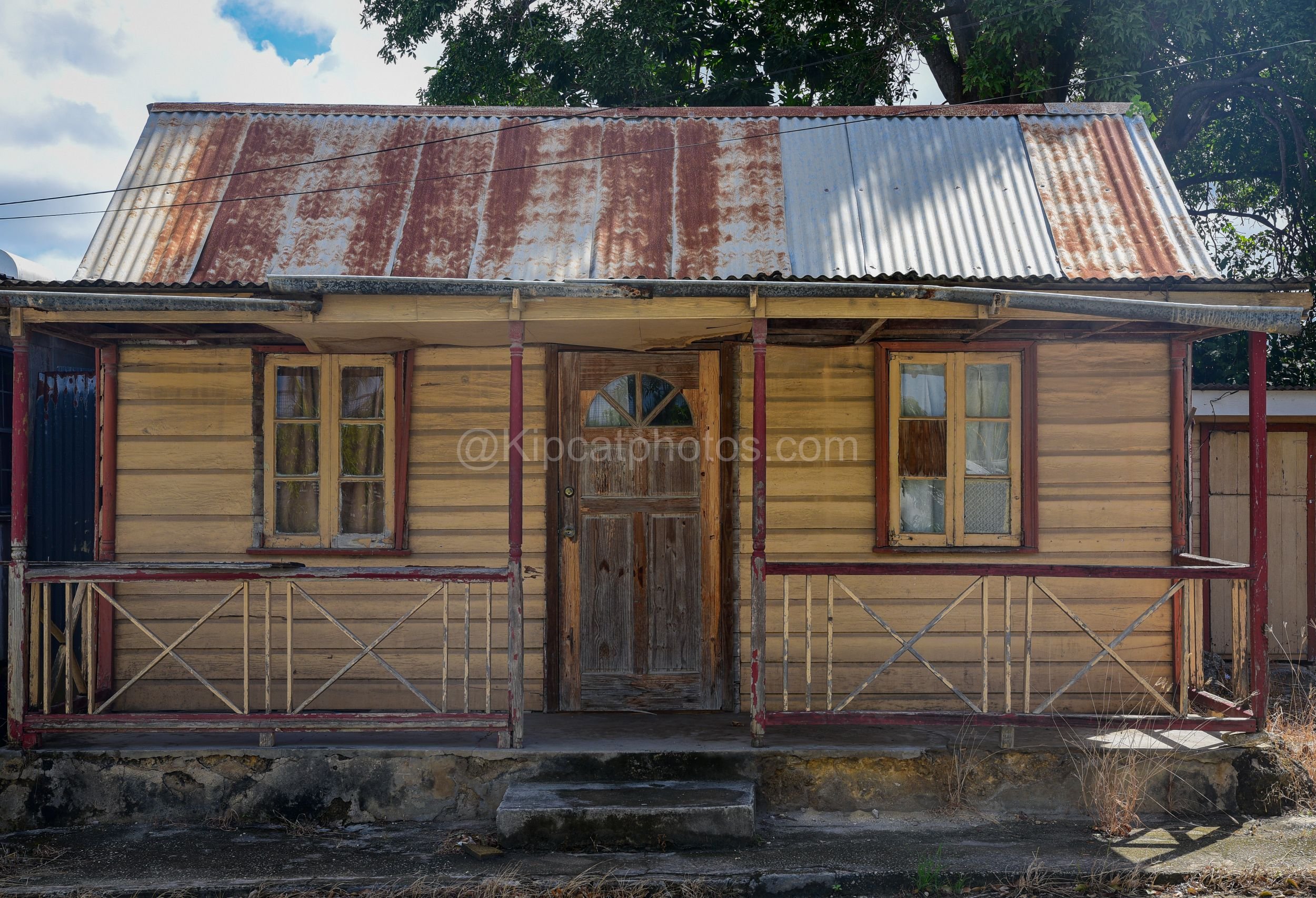 A tan chattel house in Barbados with red porch posts, a crisscross balustrade and an arched fanlight door. Kipcatphotos.com

Weathered tan clapboard and deep red porch posts frame this chattel house's arched fanlight door, its crisscross timber balus