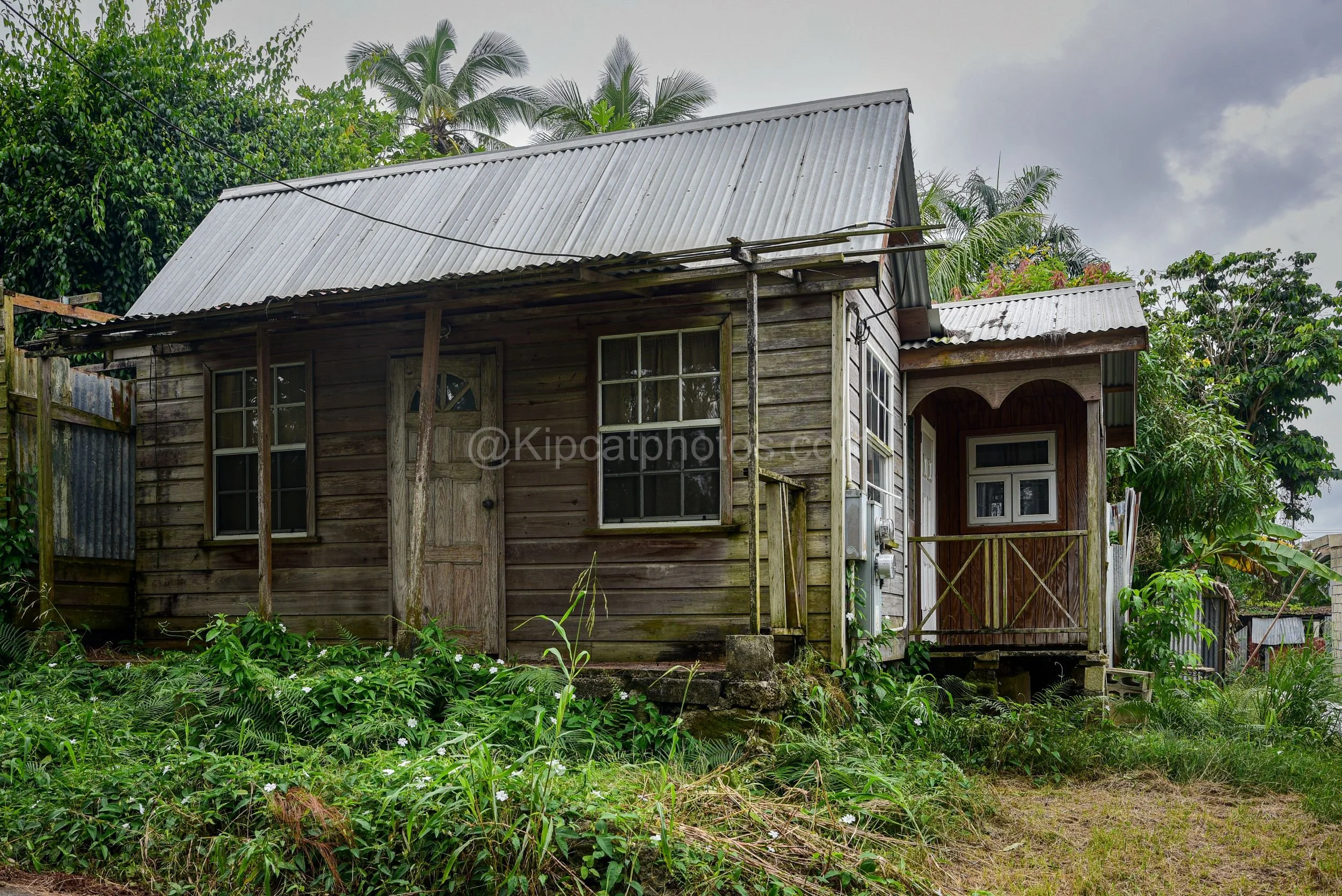 A weathered brown chattel house in Barbados with a decorative arched porch entrance and palm trees behind. Kipcatphotos.com

Rich brown timber siding and a covered porch with a decorative arched cutout give this chattel house real charm, its corrugat