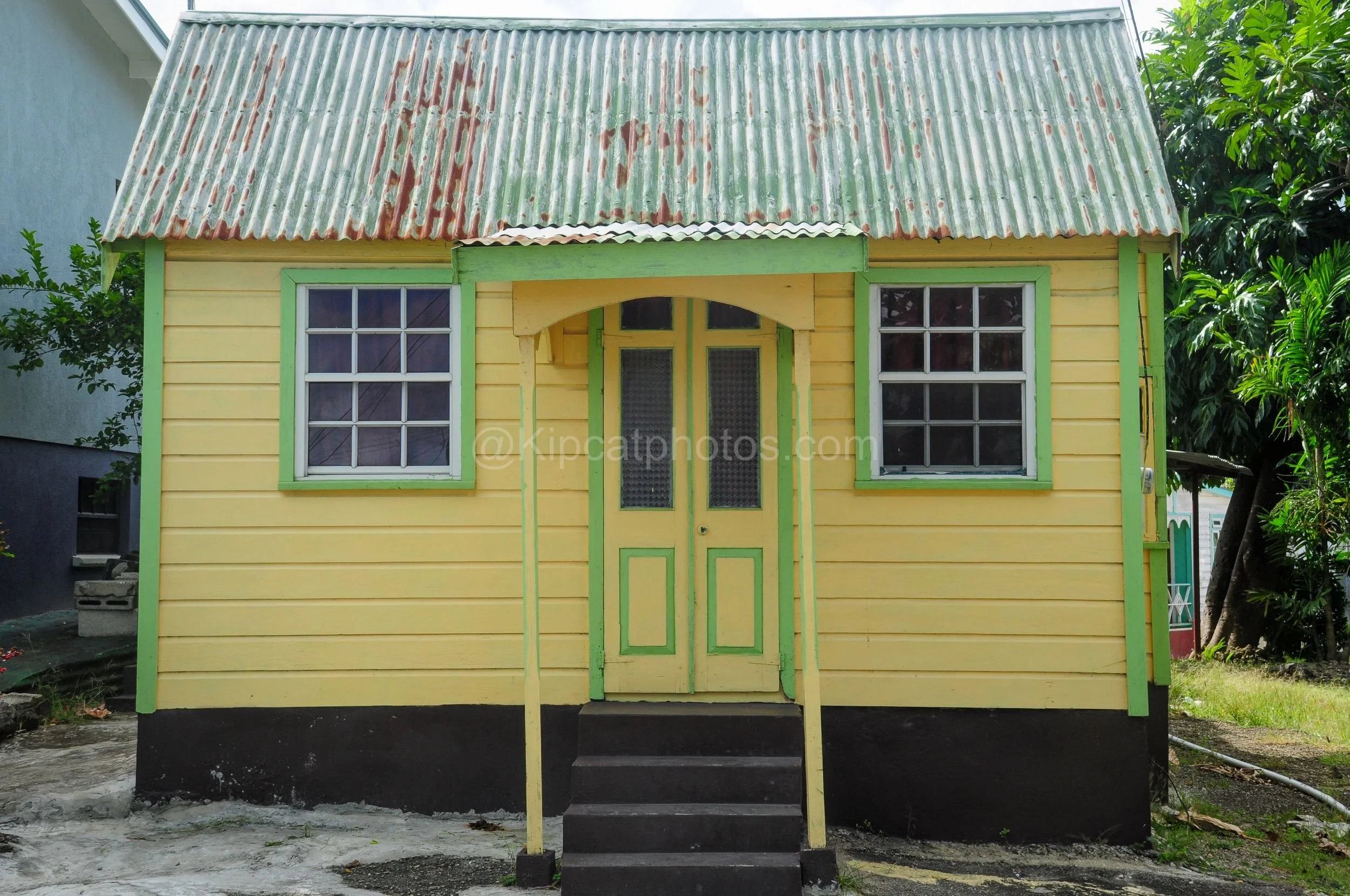 A bright yellow chattel house in Barbados with green trim and an arched porch entrance. Kipcatphotos.com

This cheerful yellow chattel house features green-trimmed windows and an arched porch overhang framing a matching green and yellow double door, 