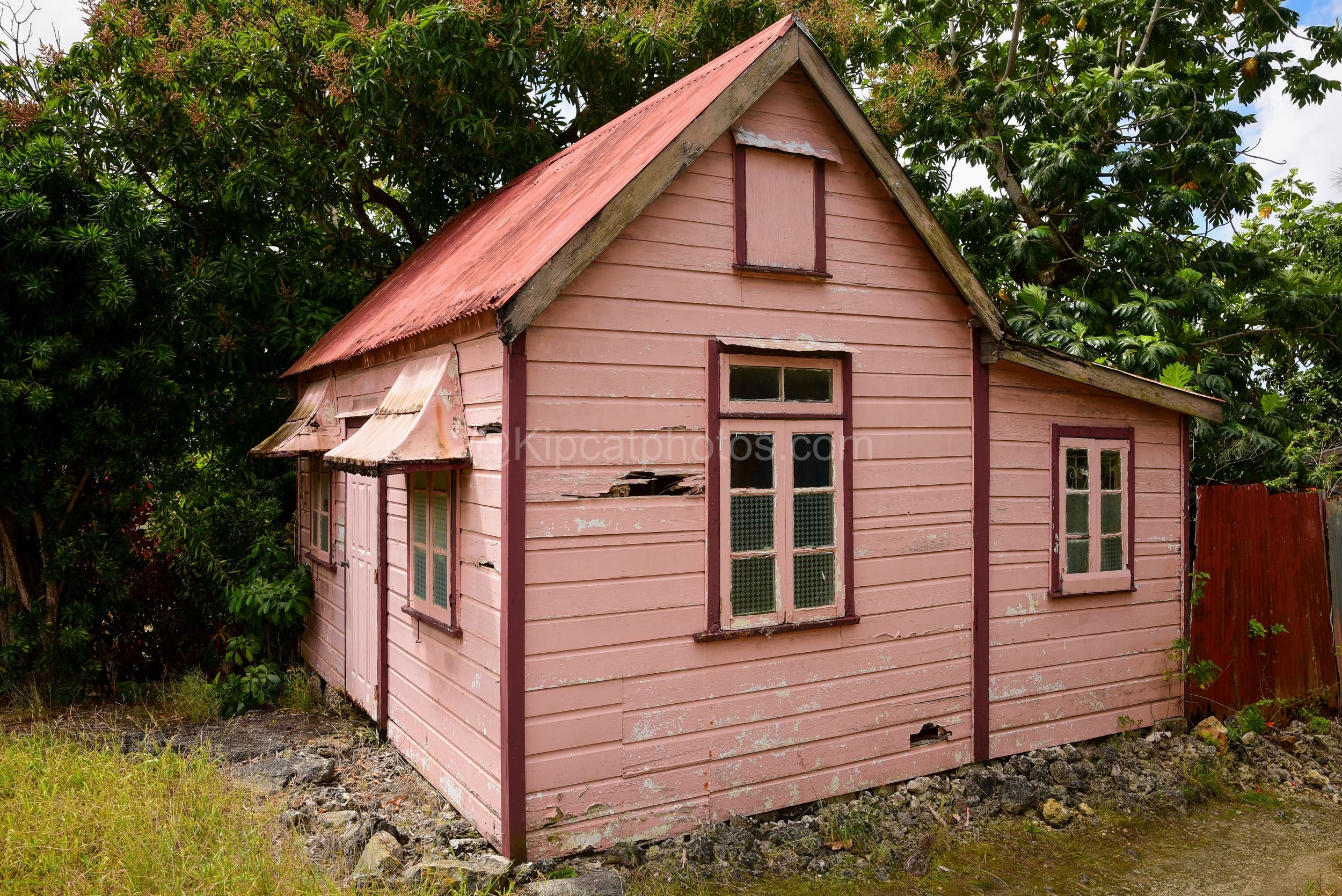 A pink chattel house in Barbados with a steep gable roof and dormer window. Kipcatphotos.com

Dusty pink paint peels from this chattel house's clapboard walls, its steep corrugated roof rising to a small dormer window, with rust-streaked window awnin