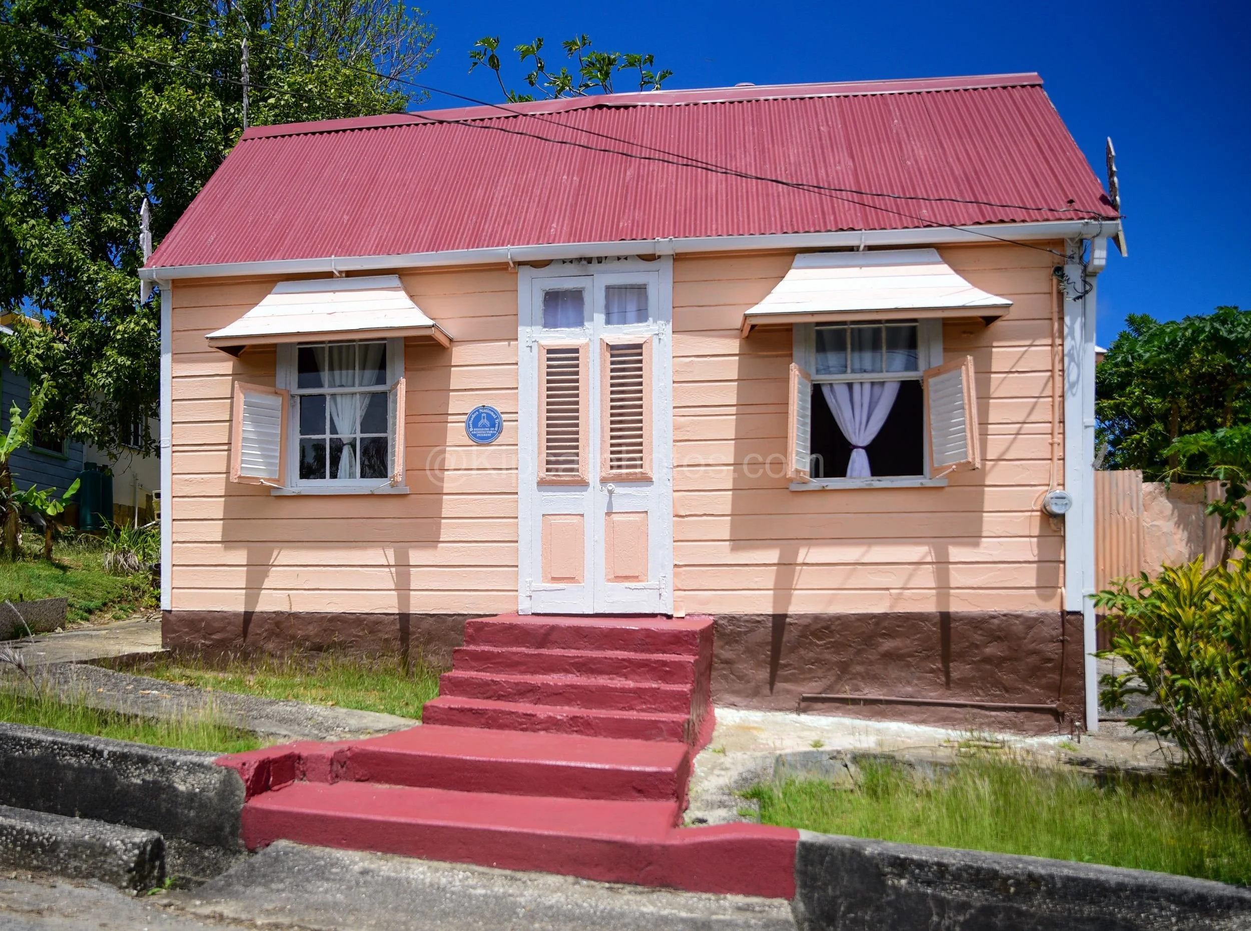 A peach-coloured chattel house in Barbados with a Barbados National Trust plaque and red painted steps. Kipcatphotos.com

Neatly restored in soft peach and white, this chattel house carries a Barbados National Trust plaque marking it as a building of