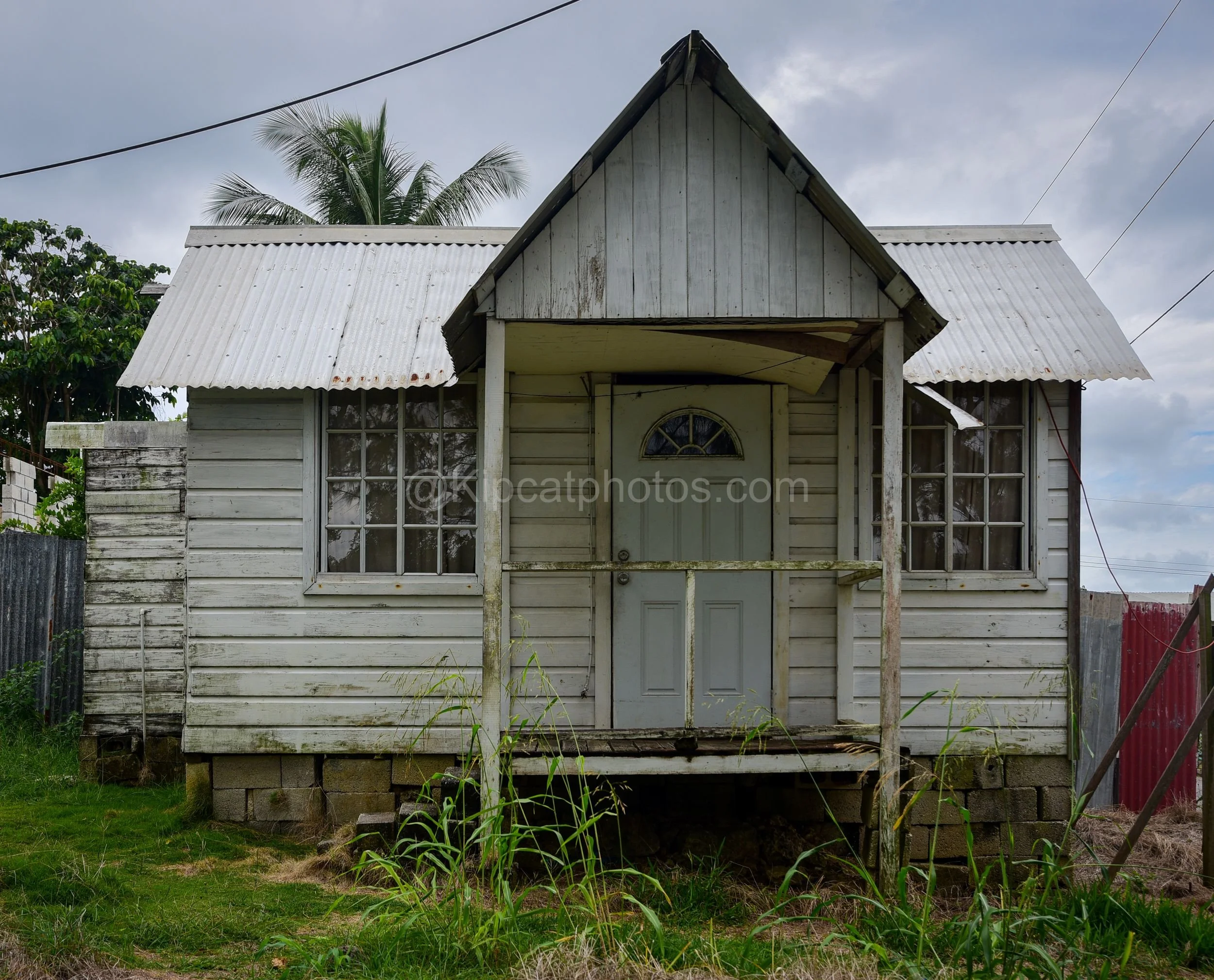 A white wooden chattel house in Barbados with a gabled porch, arched fanlight door and tin roof. Kipcatphotos.com

This white-painted chattel house features a steep gabled porch and a panelled door topped with an arched fanlight, its corrugated roof 