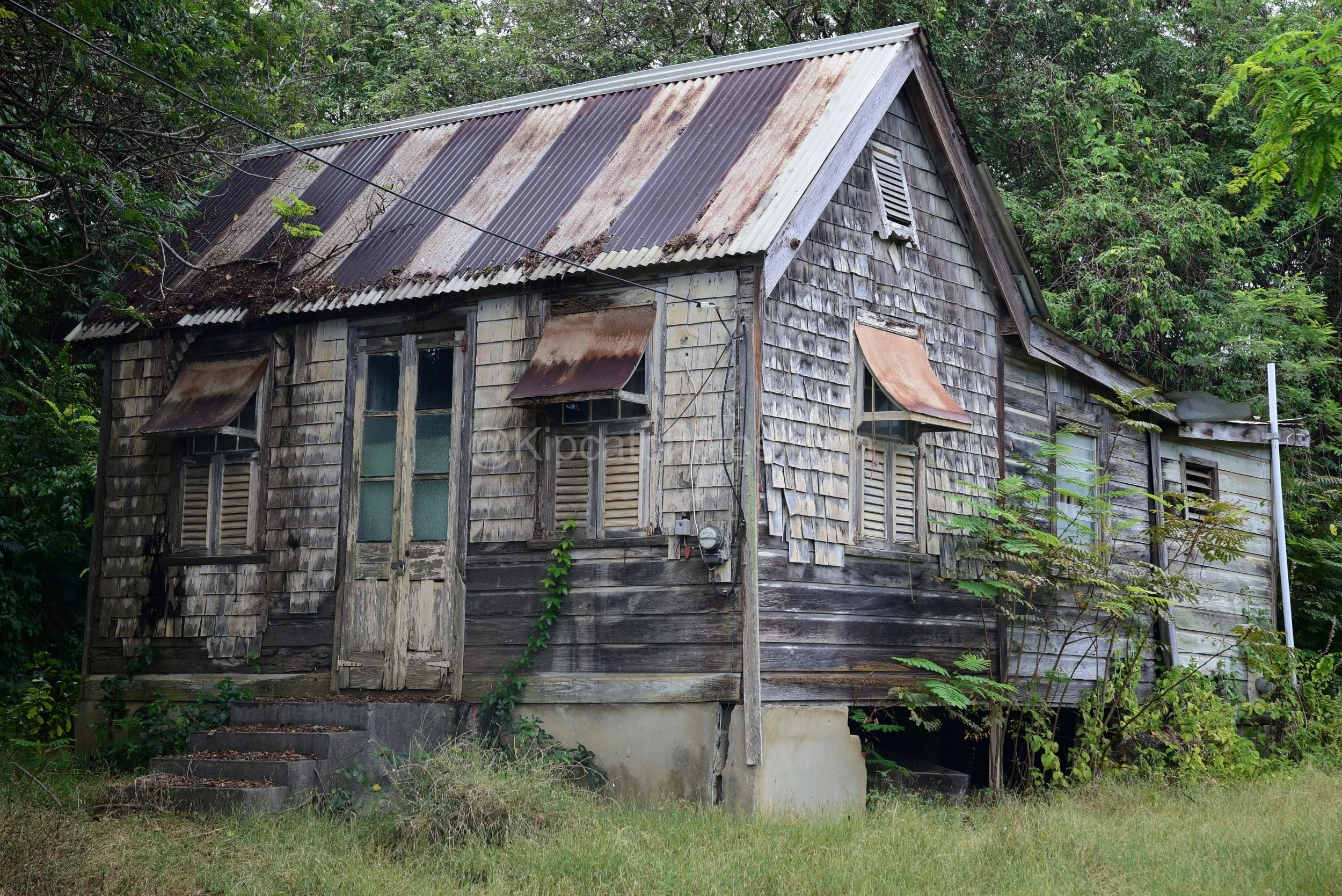 A shingle-clad chattel house in St Andrew, Barbados, with rusted window awnings and French doors. Kipcatphotos.com

Weathered wood shingles cover this chattel house in St Andrew, with rust-streaked corrugated hoods shading the windows and a pair of g