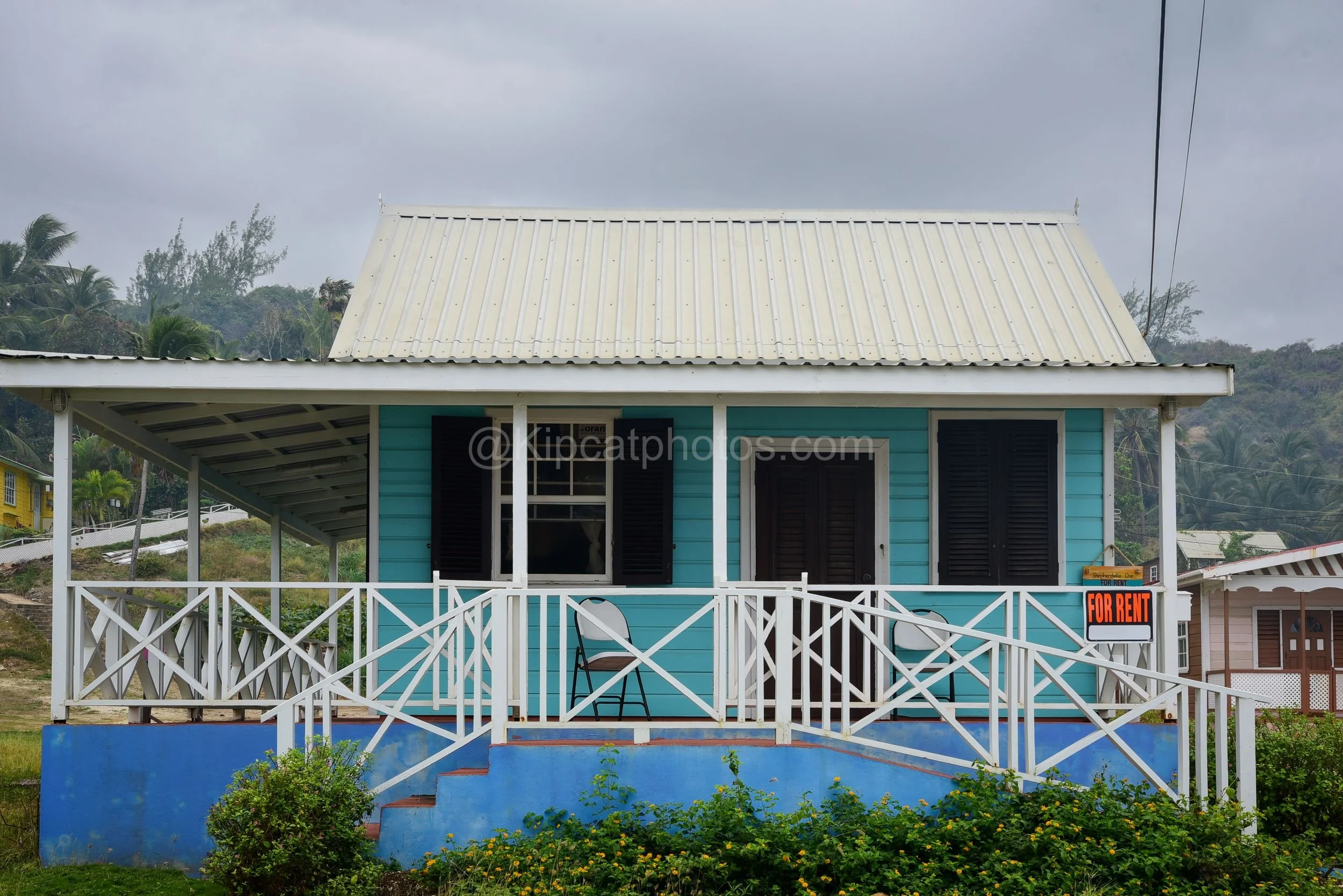 A turquoise chattel house in Barbados with a white trellis porch and a "For Rent" sign. Kipcatphotos.com

Freshly painted turquoise and white, this chattel house has a welcoming covered porch with crisscross trellis railings, folding chairs set out, 
