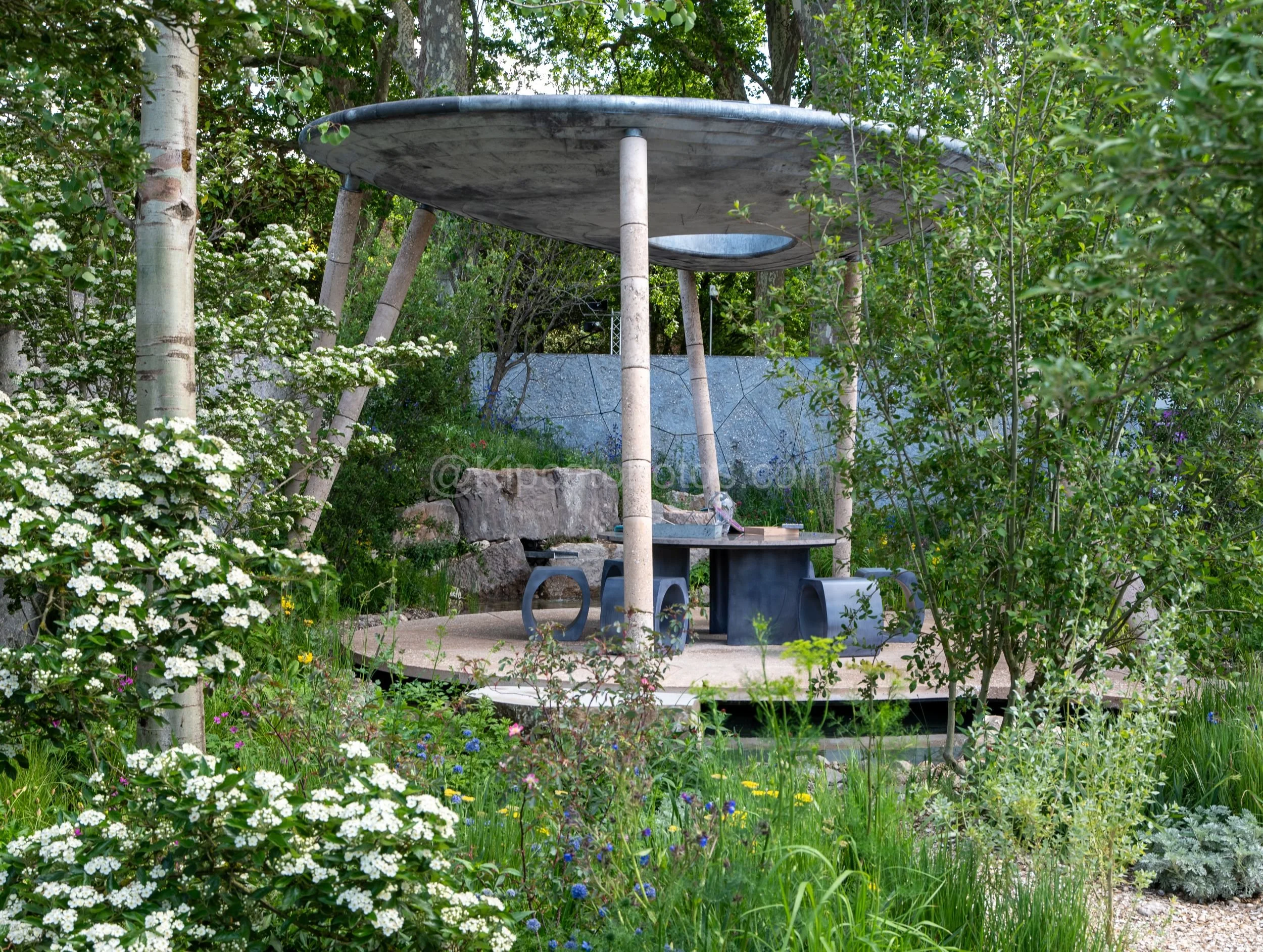 Sculptural disc-shaped canopy over a stone seating area in the Harry Holding & Alex Michaelis garden at RHS Chelsea Flower Show 2026

An UFO-like circular concrete canopy floats above a stone dining set and sculptural black stools, glimpsed through b