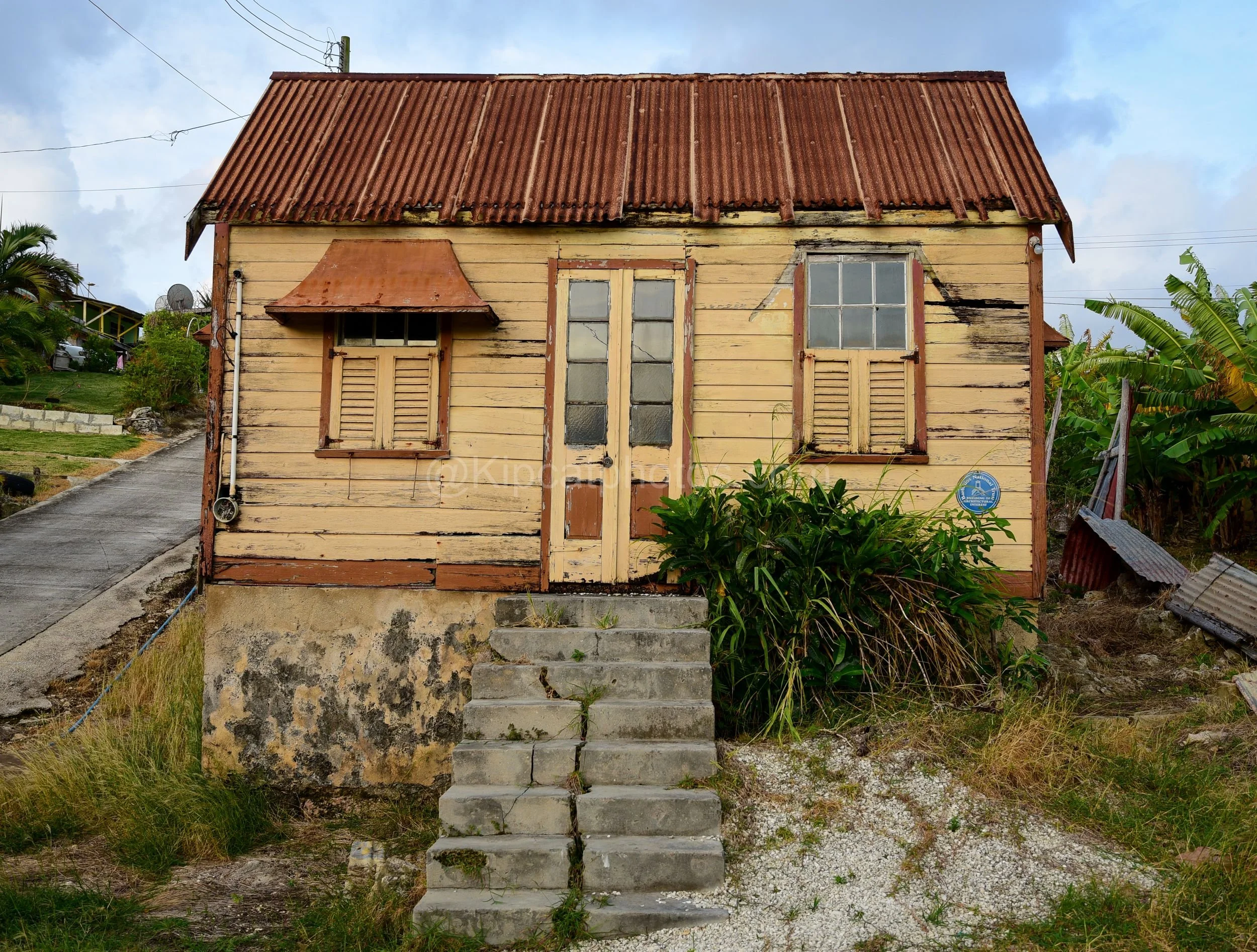 A yellow chattel house in Barbados with a Barbados National Trust plaque and a rust-streaked roof. Kipcatphotos.com

This yellow chattel house, marked with a Barbados National Trust plaque for its architectural interest, shows a rust-streaked corruga