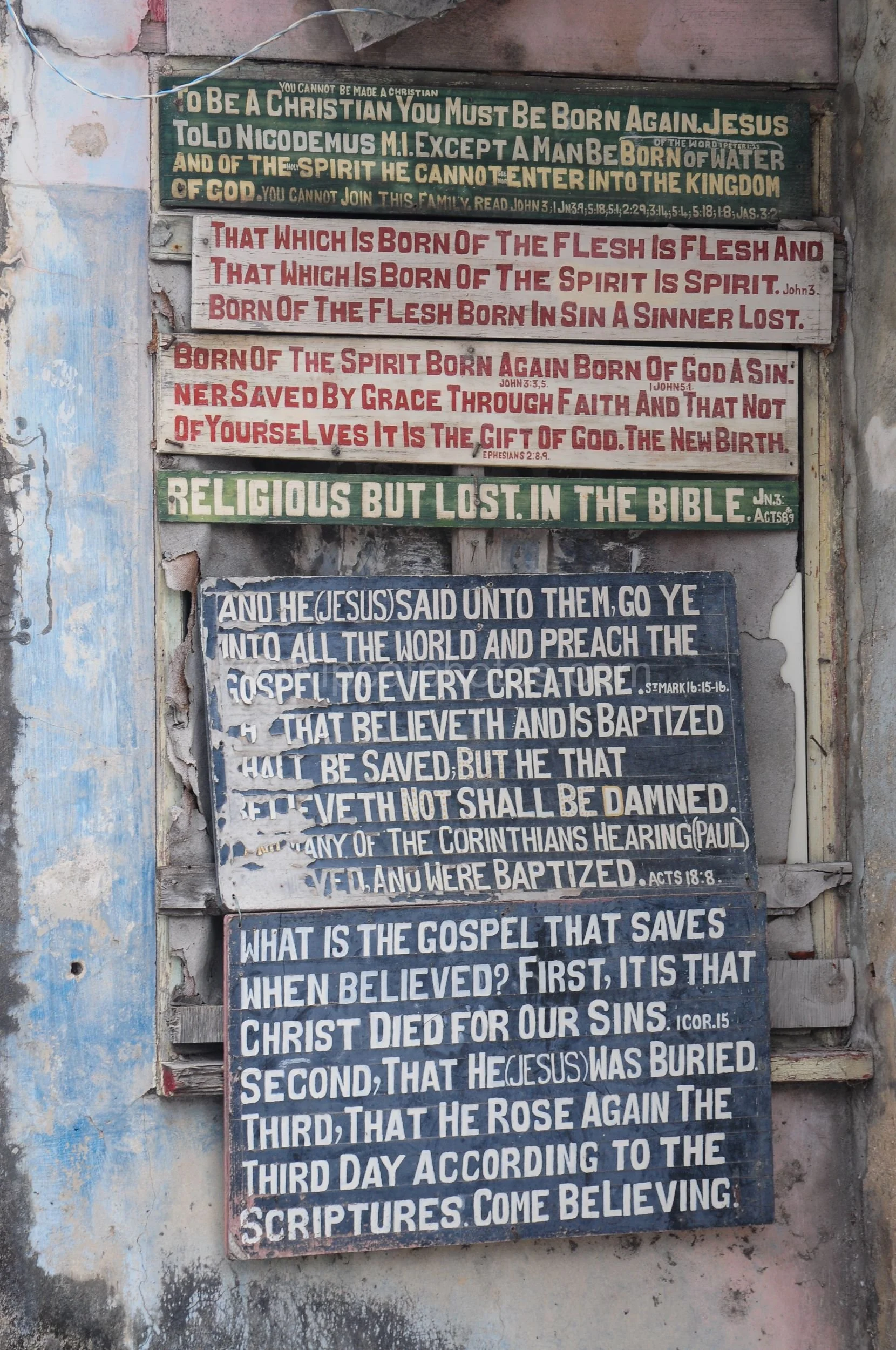 A close-up of hand-painted scripture signs fixed to a weathered wall in Barbados. Kipcatphotos.com

Stacked hand-lettered signs quote Bible verses on faded green, white and blue boards, nailed to a crumbling plastered wall as part of an informal road