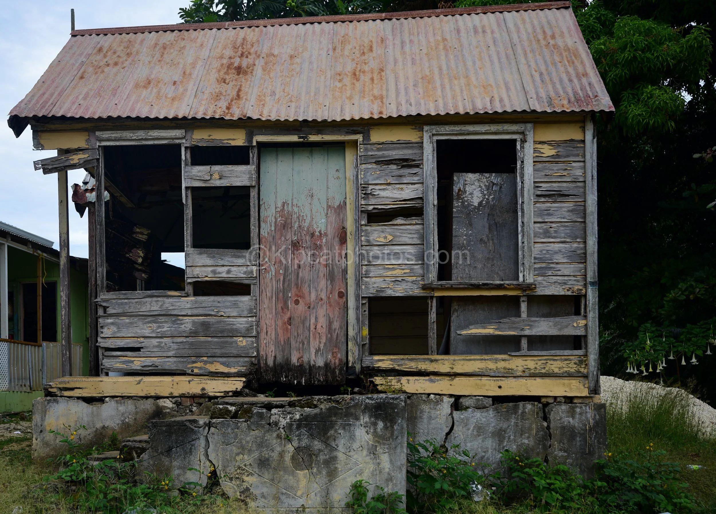 A dilapidated wooden chattel house in Barbados with a weathered blue door and a boarded window. Kipcatphotos.com

Grey, sun-bleached boards and a flaking blue door mark this abandoned chattel house, its window boarded over and its cracked concrete ba