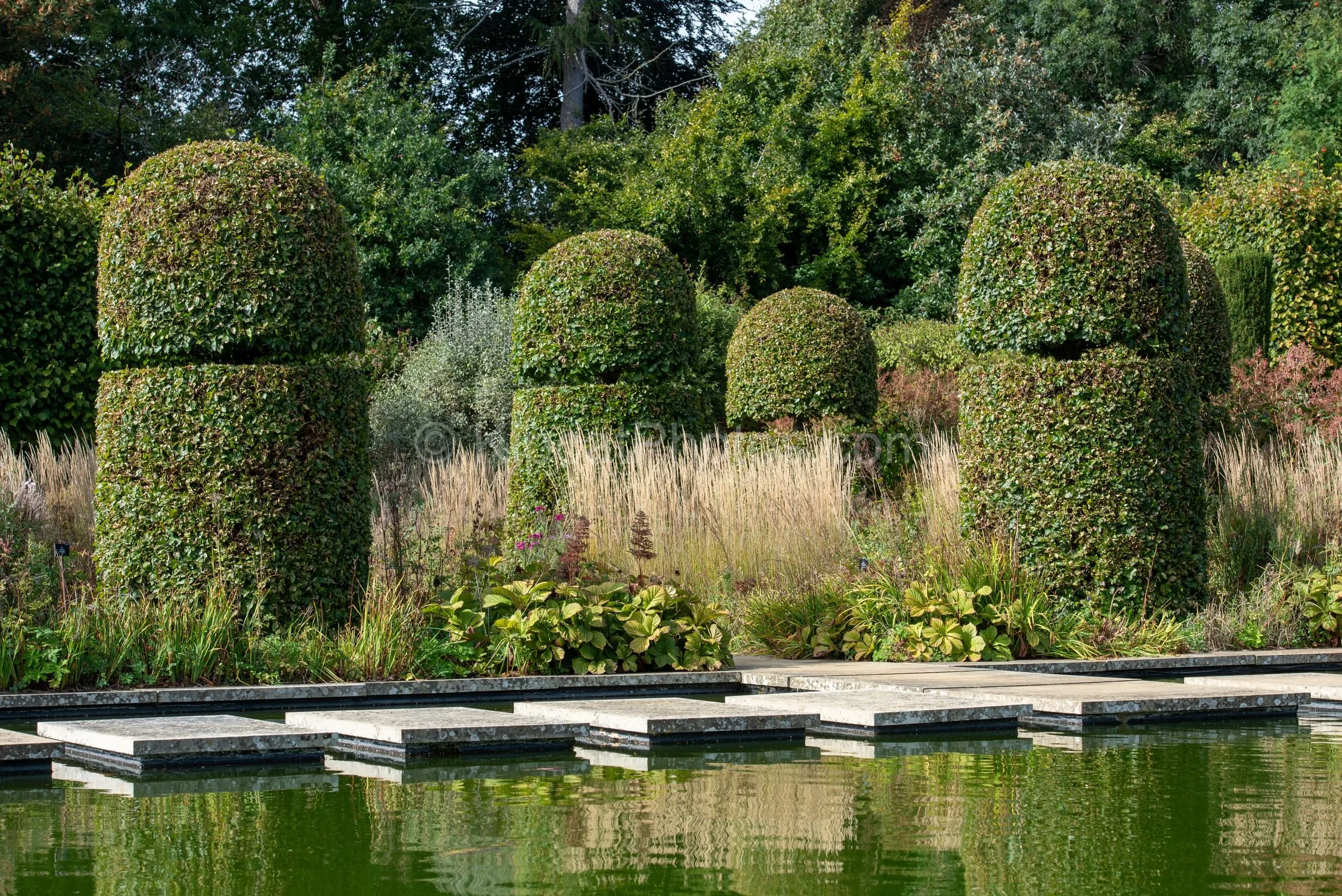 Stone stepping stones cross the formal canal pool at Broughton Grange, Oxfordshire, with large hornbeam dome topiary columns and golden ornamental grasses reflected in the still water, and the arboretum trees beyond. KipcatPhotos.com.

This view acro