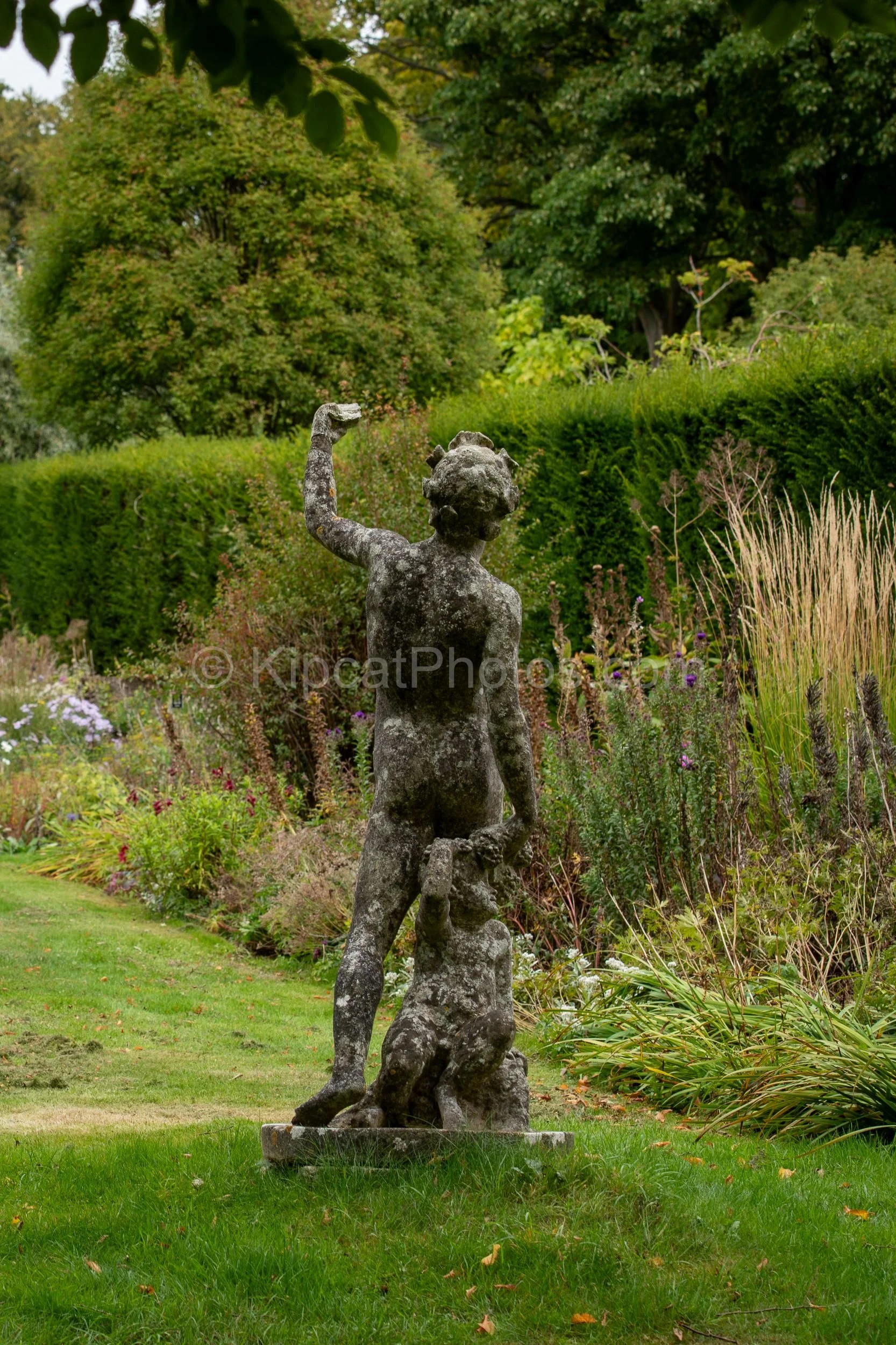 A weathered stone figure sculpture stands beside the double herbaceous borders at Broughton Grange, Oxfordshire, surrounded by ornamental grasses, asters and late perennials with a yew hedge backdrop. KipcatPhotos.com.

This lichen-covered stone figu
