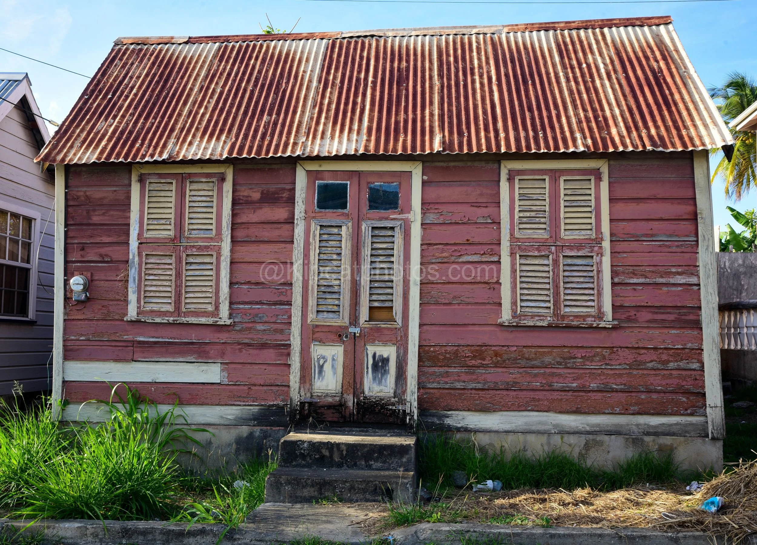 A dusty pink chattel house in Barbados with cream-trimmed louvred shutters and a panelled door. Kipcatphotos.com

Faded dusty pink paint peels from this chattel house's timber walls, its cream-trimmed louvred shutters and glass-paned door showing the