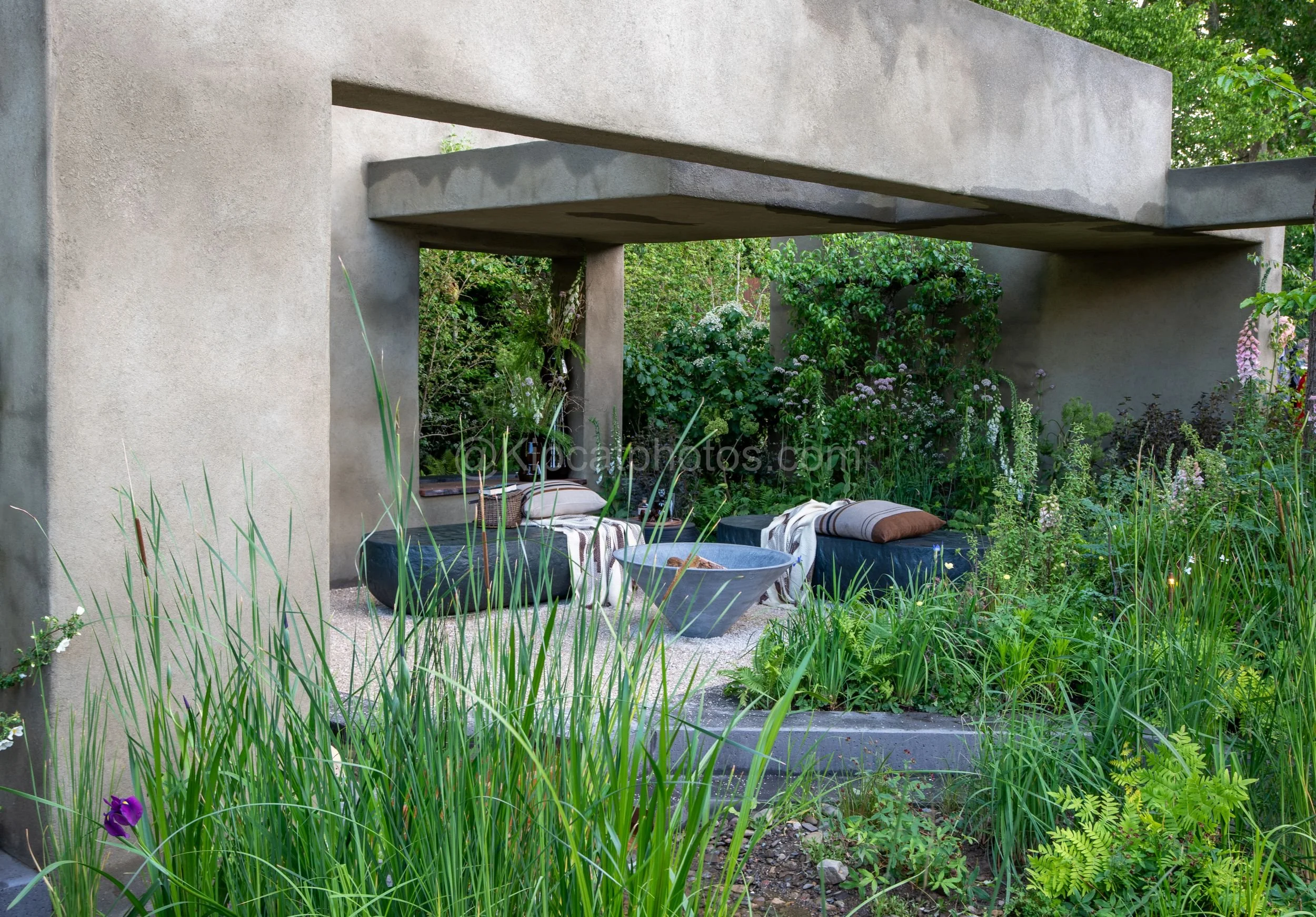 Concrete pergola framing a sunken seating area in the Baz Grainger garden at RHS Chelsea Flower Show 2026

A view through a concrete pergola towards a sunken gravel seating area with sculptural stone benches and cushions, framed by reeds and iris, in