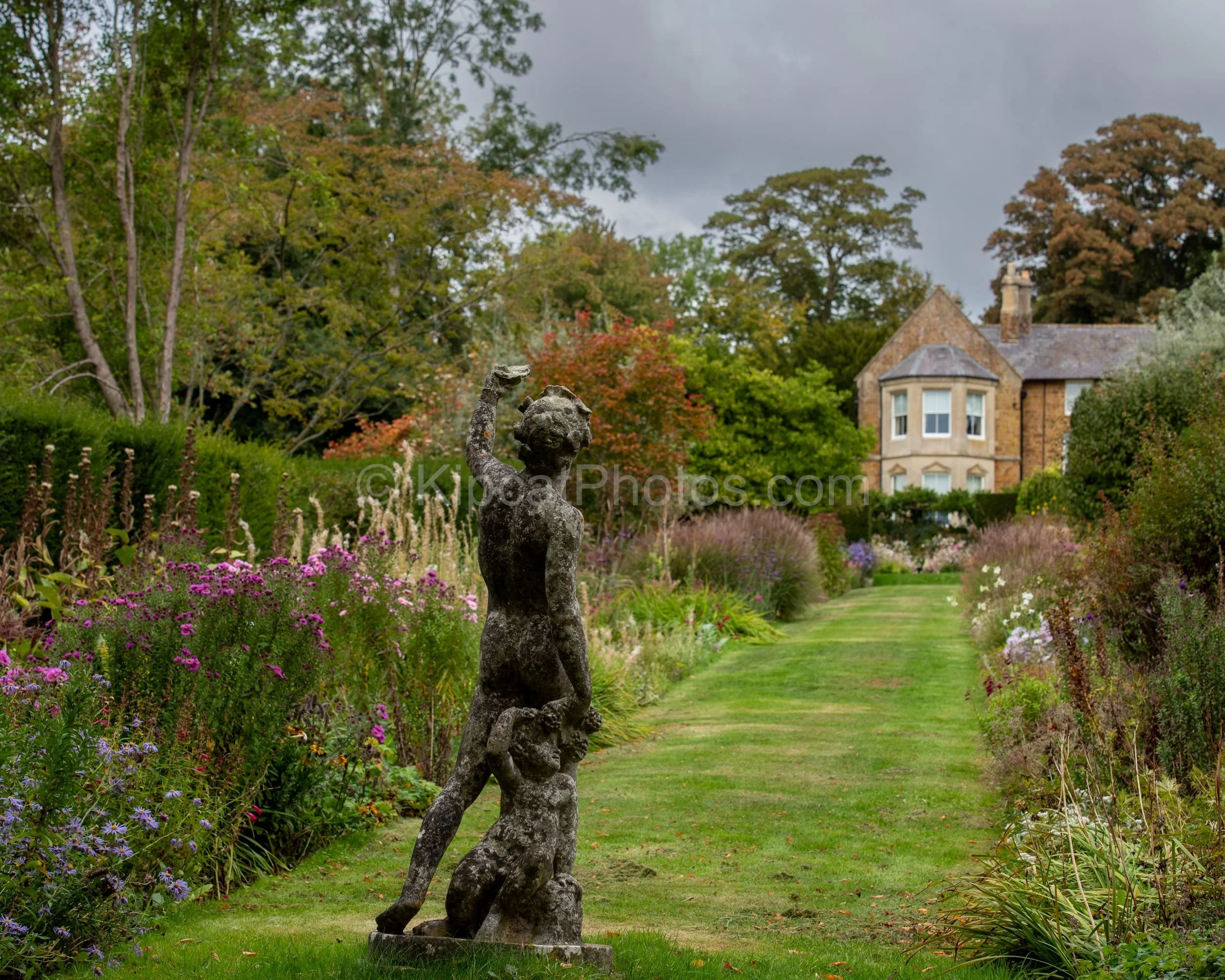 A weathered stone figure stands at the end of the double borders at Broughton Grange, Oxfordshire, with pink asters, cosmos and ornamental grasses flanking a grass path leading back towards the honey-stone manor house and autumn trees. KipcatPhotos.c