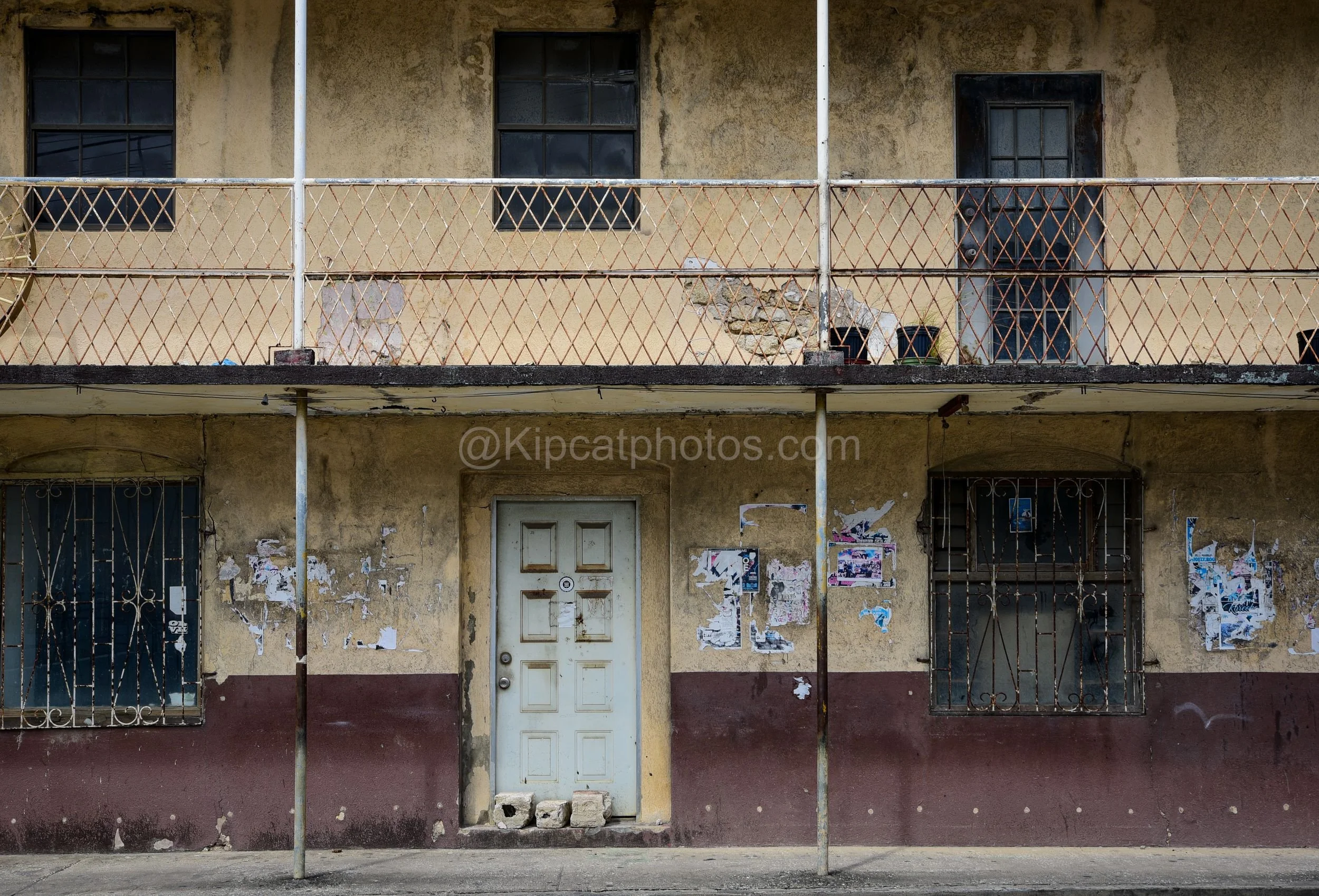 A faded two-storey building in Speightstown, Barbados, with a diamond-lattice balcony railing and peeling ochre plaster. Kipcatphotos.com

Ochre and maroon paint peels from this Speightstown building's plastered facade, its upper balcony enclosed by 