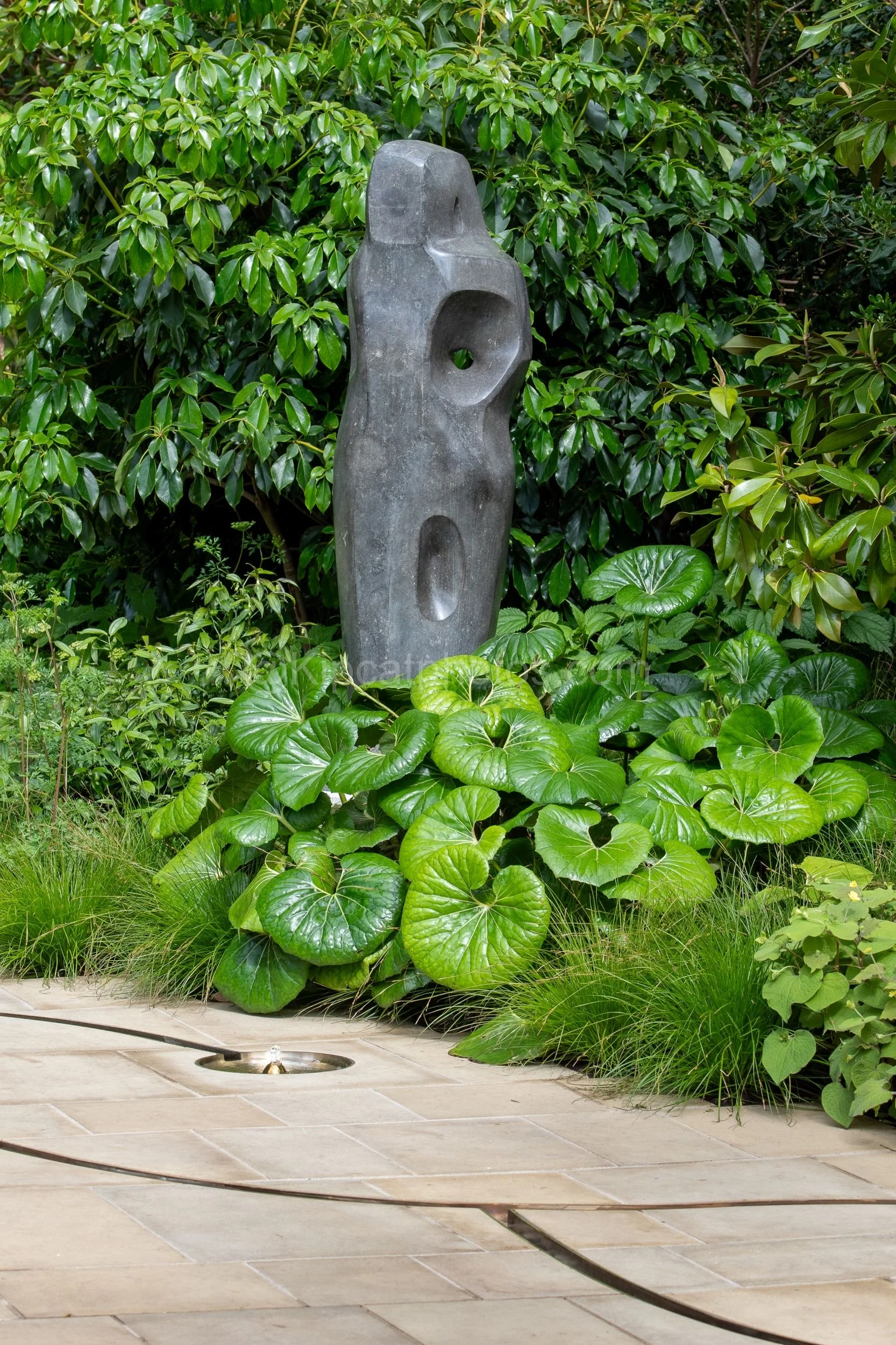 Tall abstract stone sculpture by Barbara Hepworth rising above glossy green foliage in the Tom Stuart-Smith garden at RHS Chelsea Flower Show 2026

A tall abstract carved stone sculpture by Barbara Hepworth with rounded openings stands above a bed of