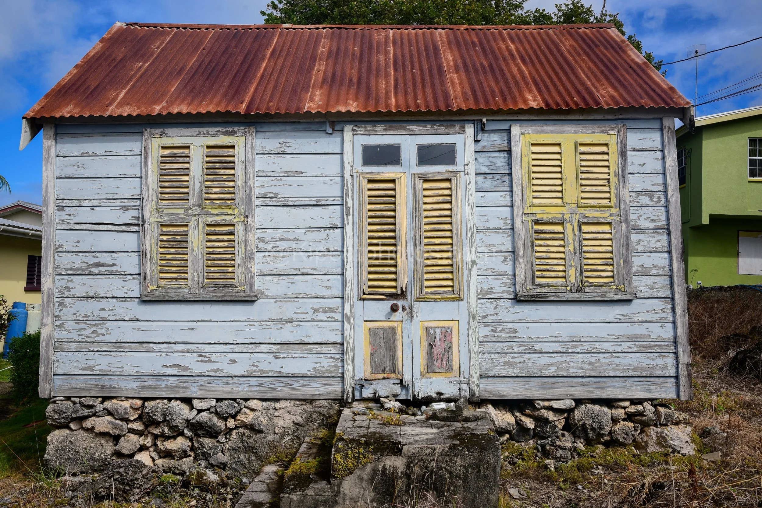 A pale blue chattel house in Barbados with yellow louvred shutters and a rusted red roof. Kipcatphotos.com

Flaking pale blue paint and cheerful yellow louvred shutters give this chattel house real character, its double door and window hoods weathere