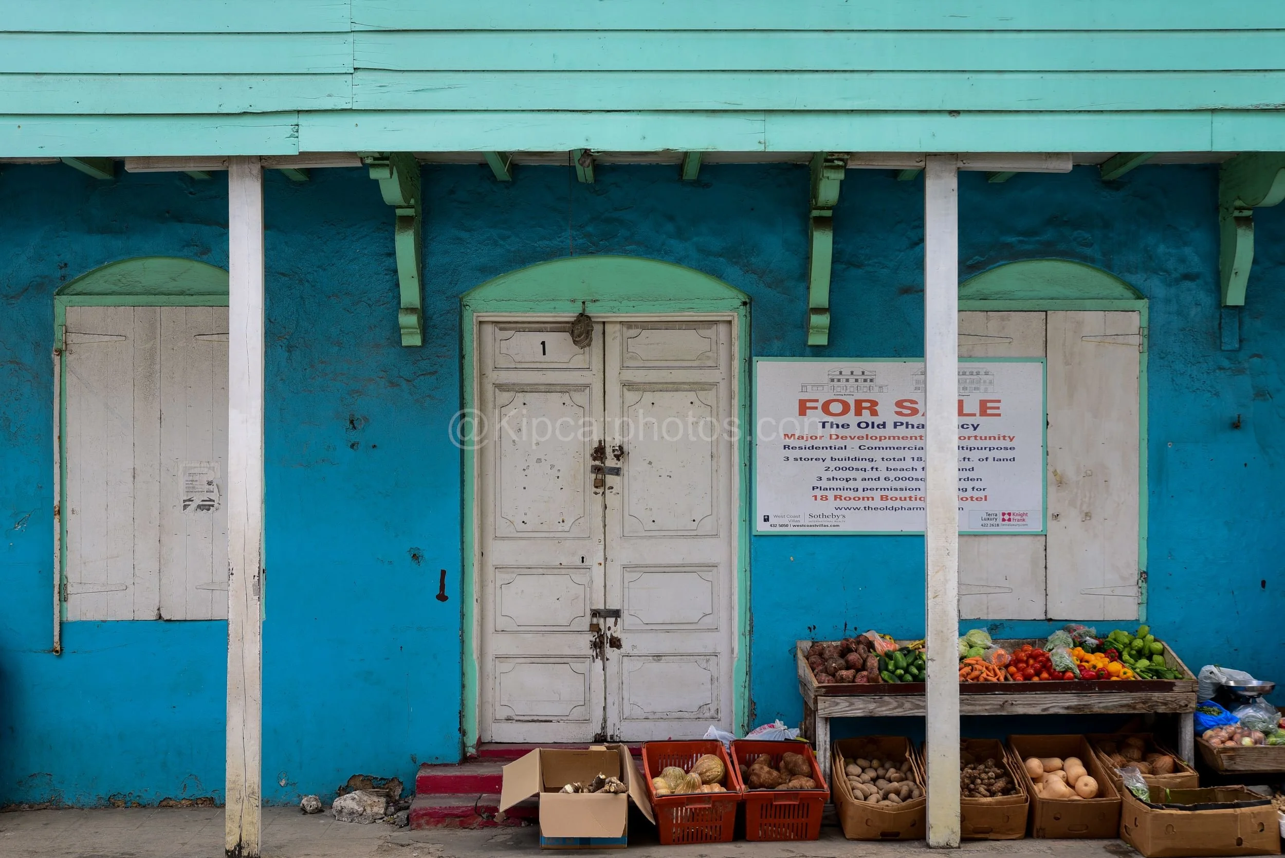 A turquoise building in Barbados with white doors and shutters, a "For Sale" sign, and a vegetable stall out front. Kipcatphotos.com

Weathered white doors and shutters stand out against this turquoise building's textured walls, a "For Sale" sign adv