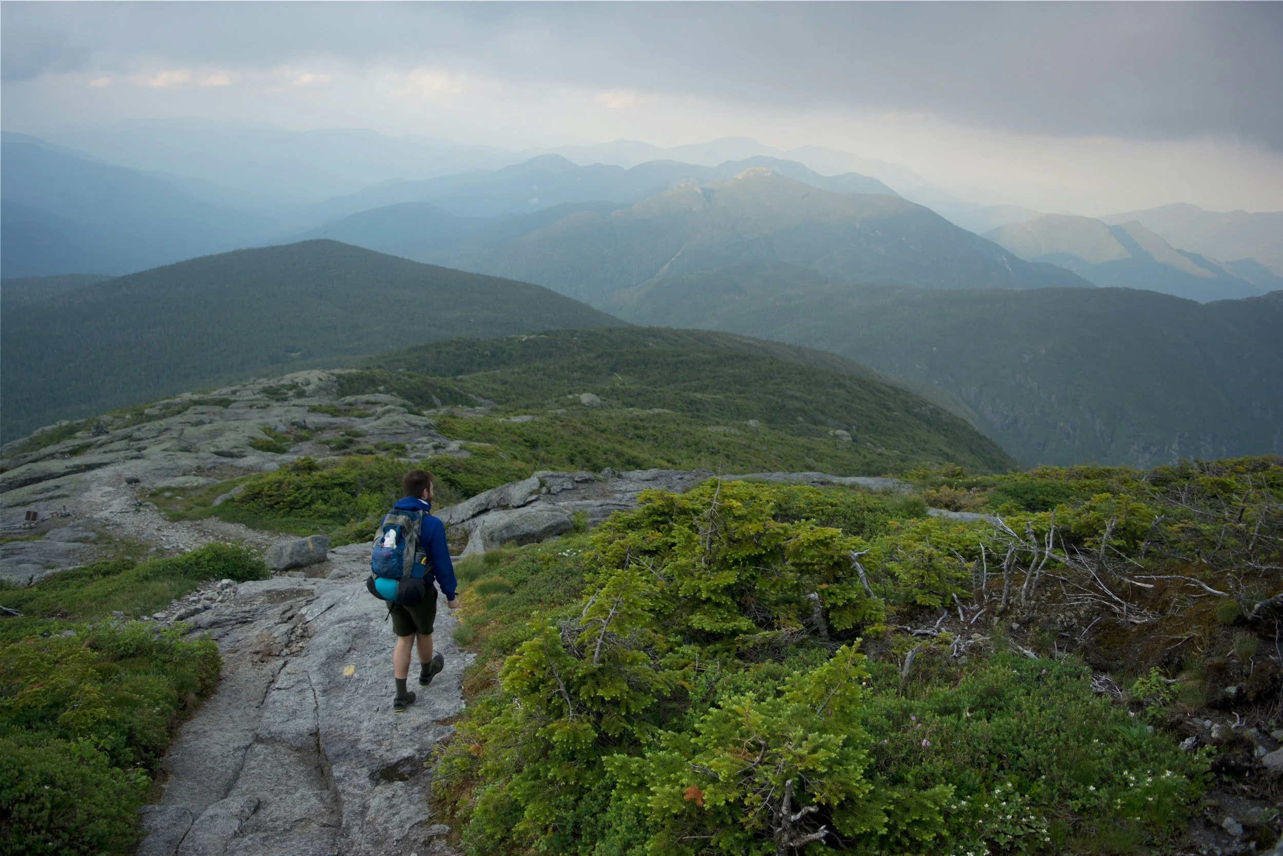 backpacker walking on a rocky trail overlooking multiple mountains in the distance