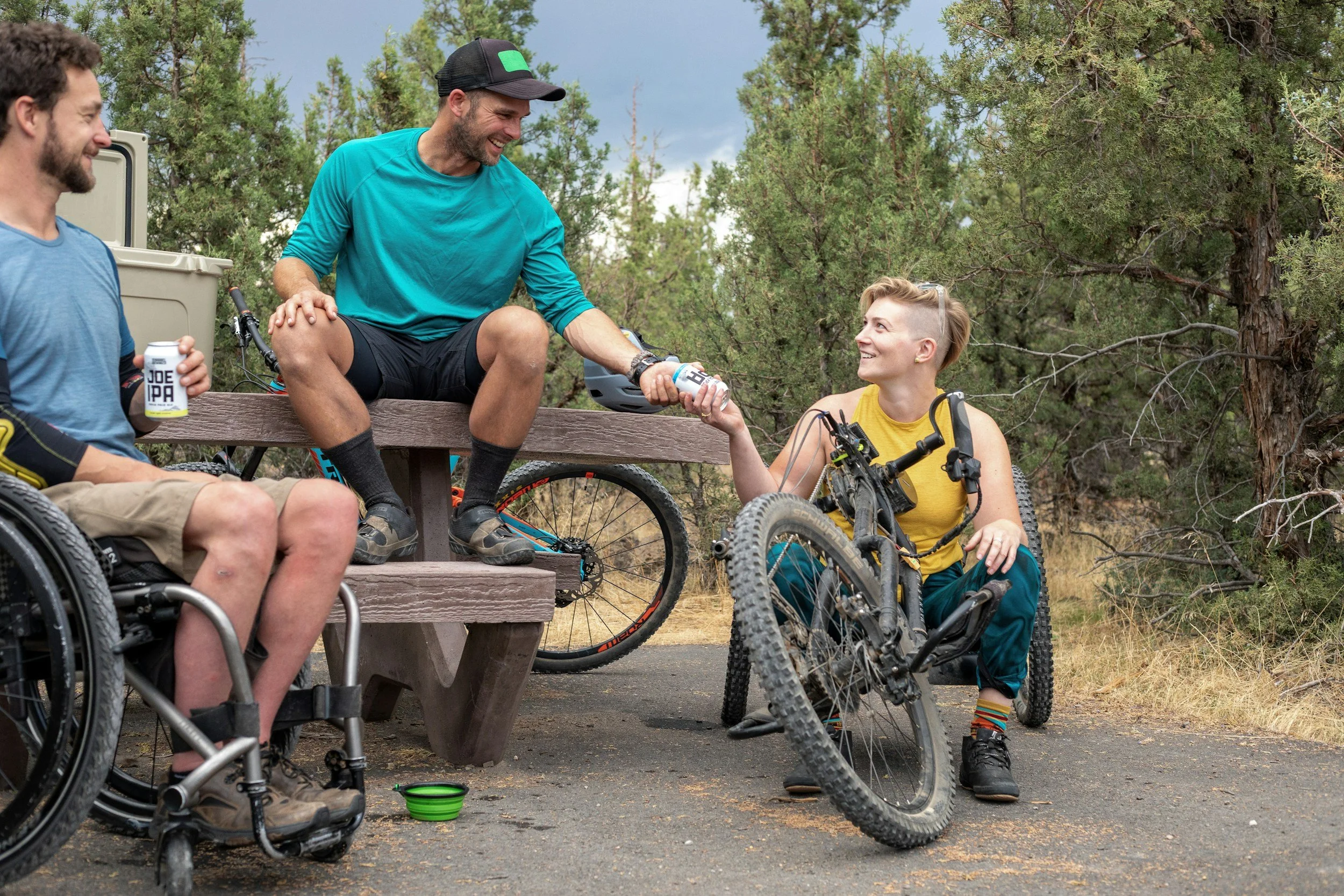 Three people at a campground sharing a beer, one in a wheelchair, one sitting on the table, and one on an adaptive bike