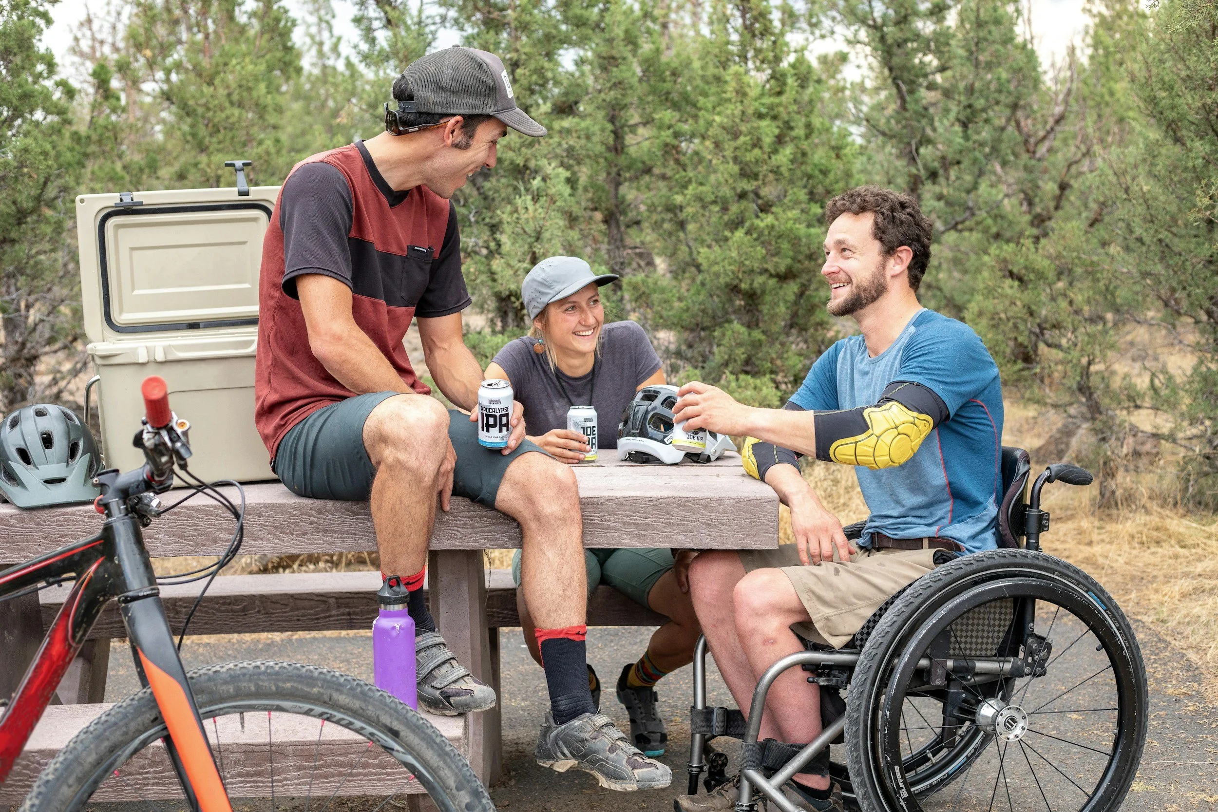 three people at a picnic table smiling drinking beer with one person in a wheelchair