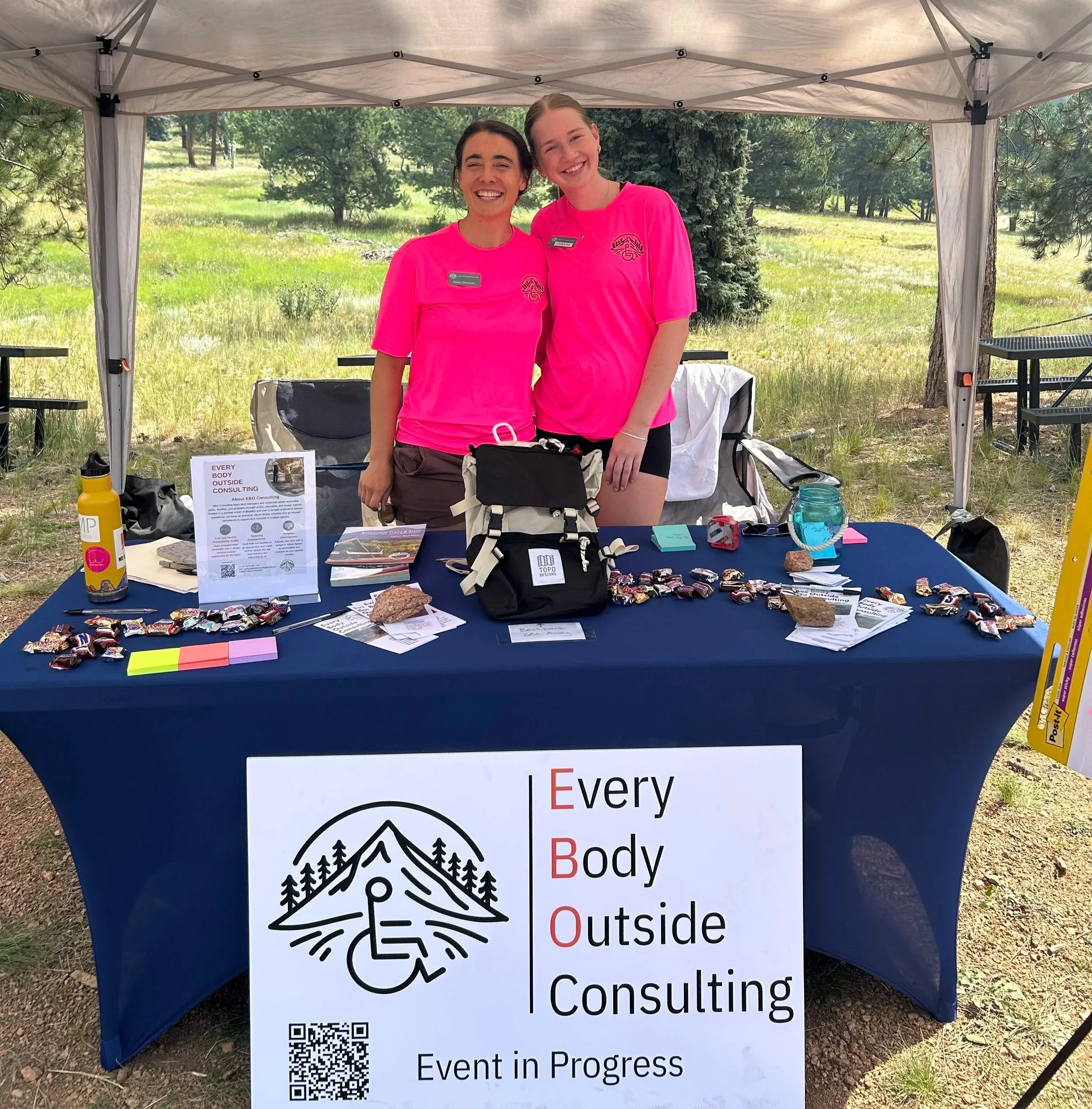 Myself and Lauren in bright pink shirts standing behind our table with a big white sign "Every Body Outside Consulting"