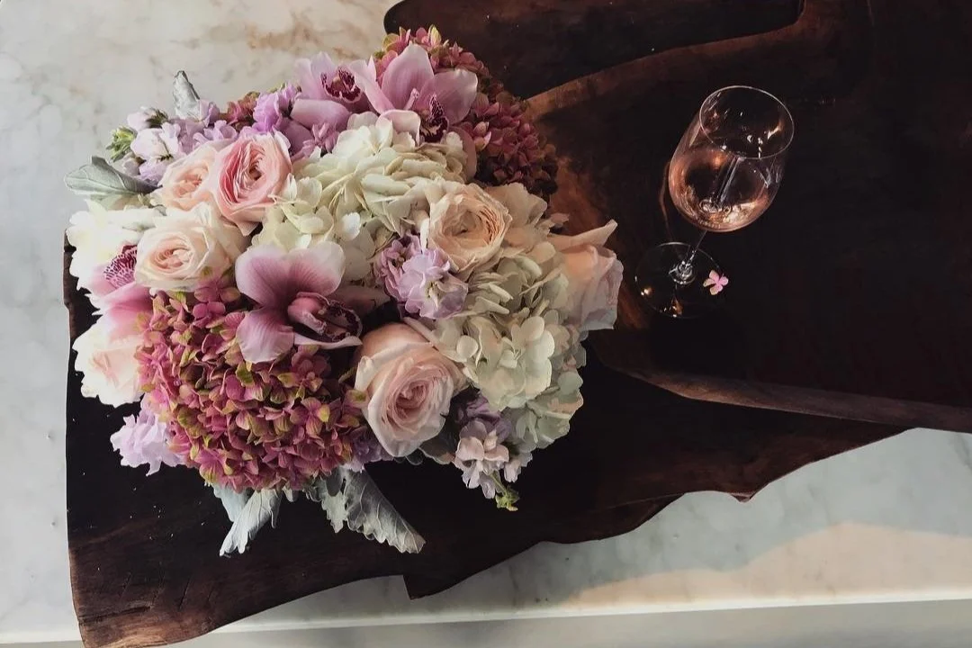 A bouquet of pink, white, and purple flowers with a glass of rosé wine on a dark wooden tray.
