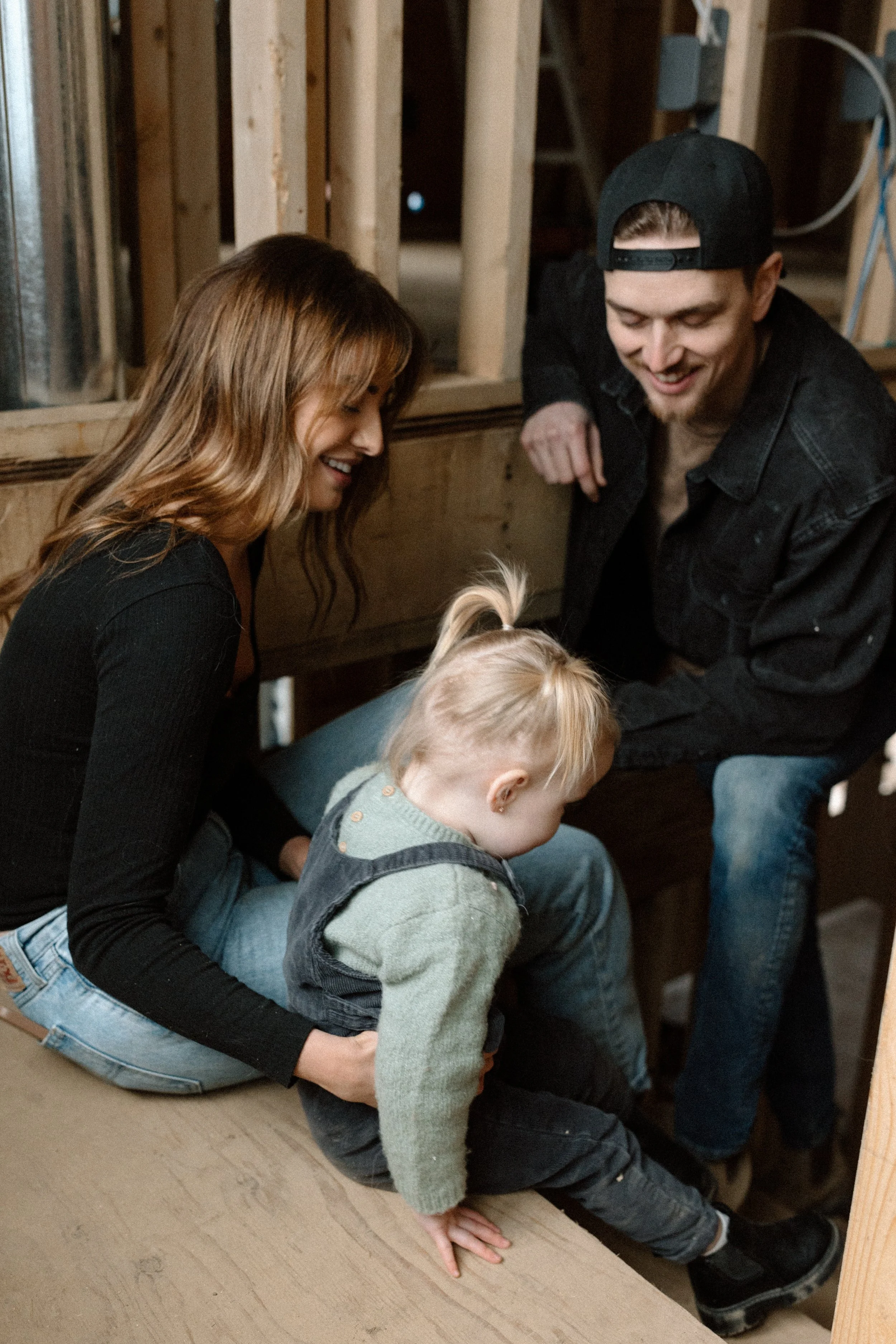 A woman, a man, and two children are sitting on a wooden floor in a partially constructed room with exposed wooden studs and electrical wiring. The woman has long, wavy hair and is wearing a black long-sleeved top and jeans. The man is wearing a black baseball cap backward, a black jacket, and jeans. The children have blonde hair; one is sitting on the floor with a hand on the wooden surface, and the other child with long hair is partially visible.