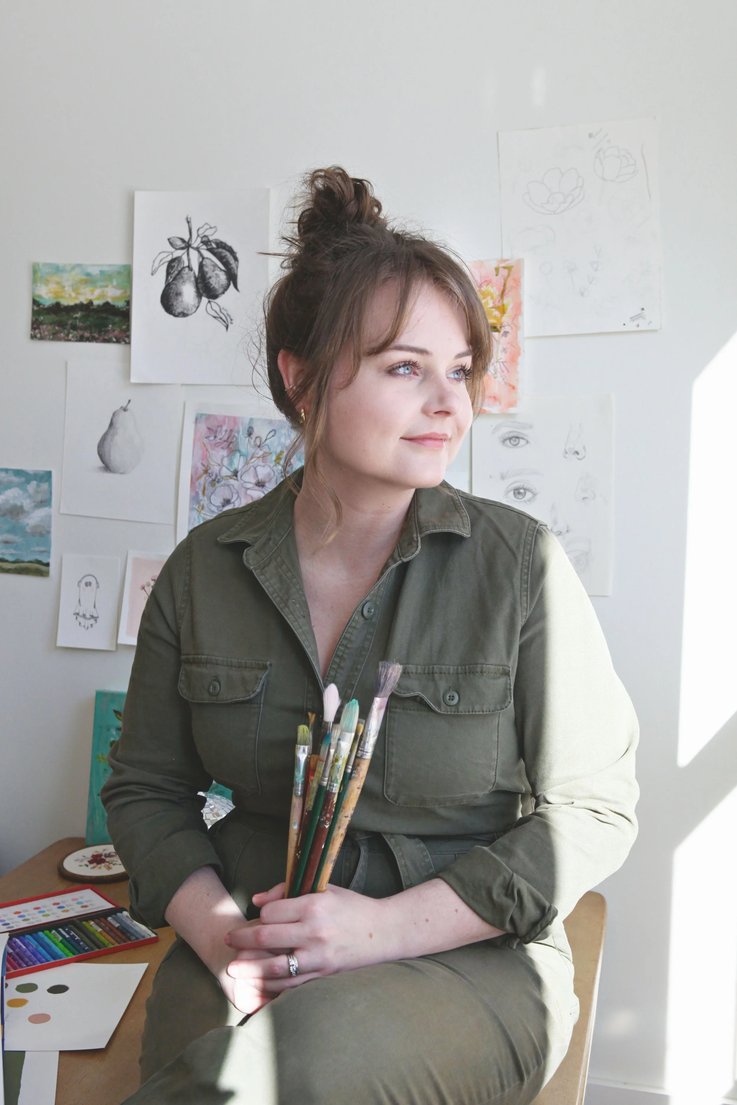 woman artist sitting in art studio looking out window holding paint brushes