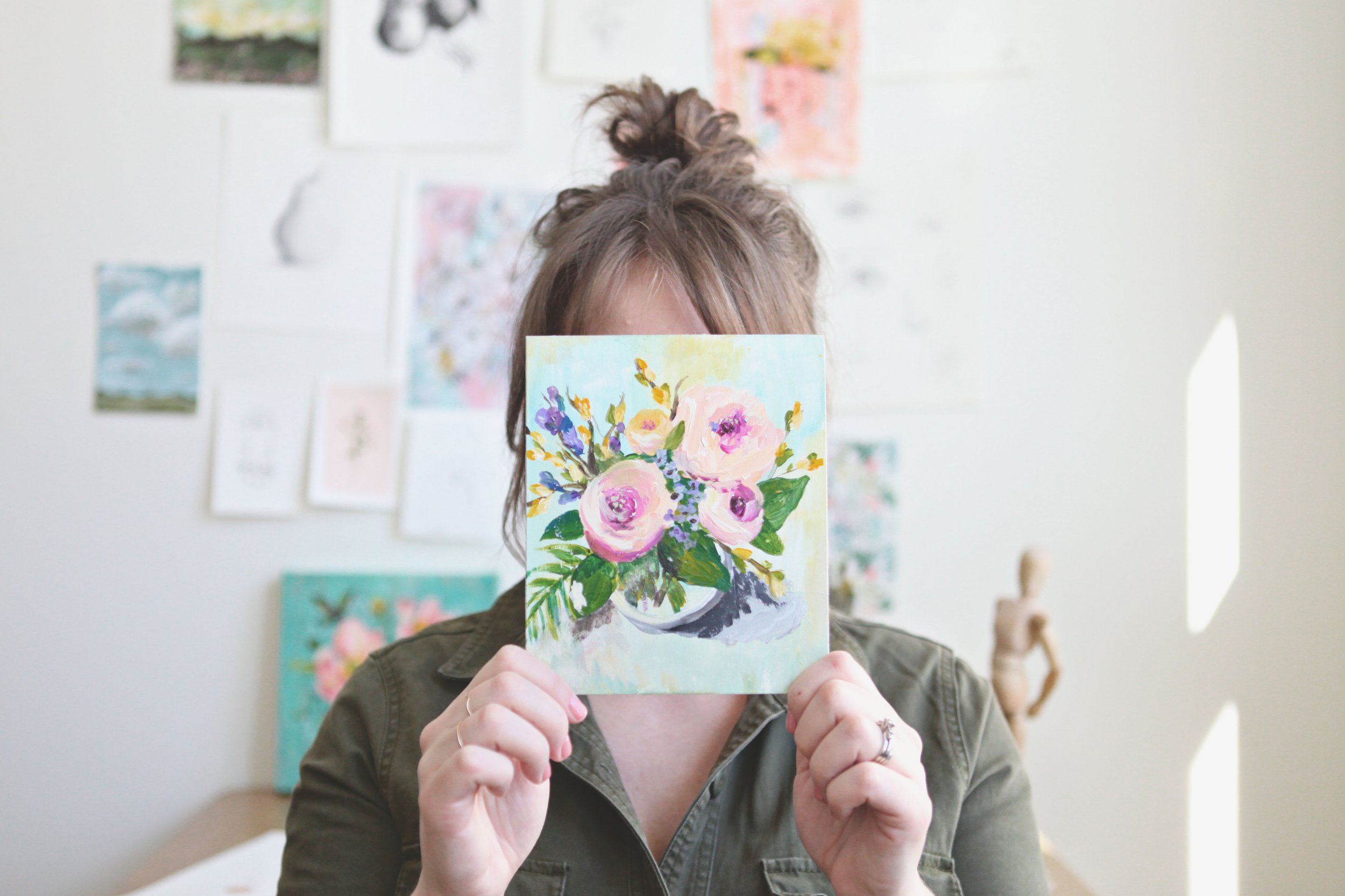 artist holding floral painting covering face in front of art studio wall