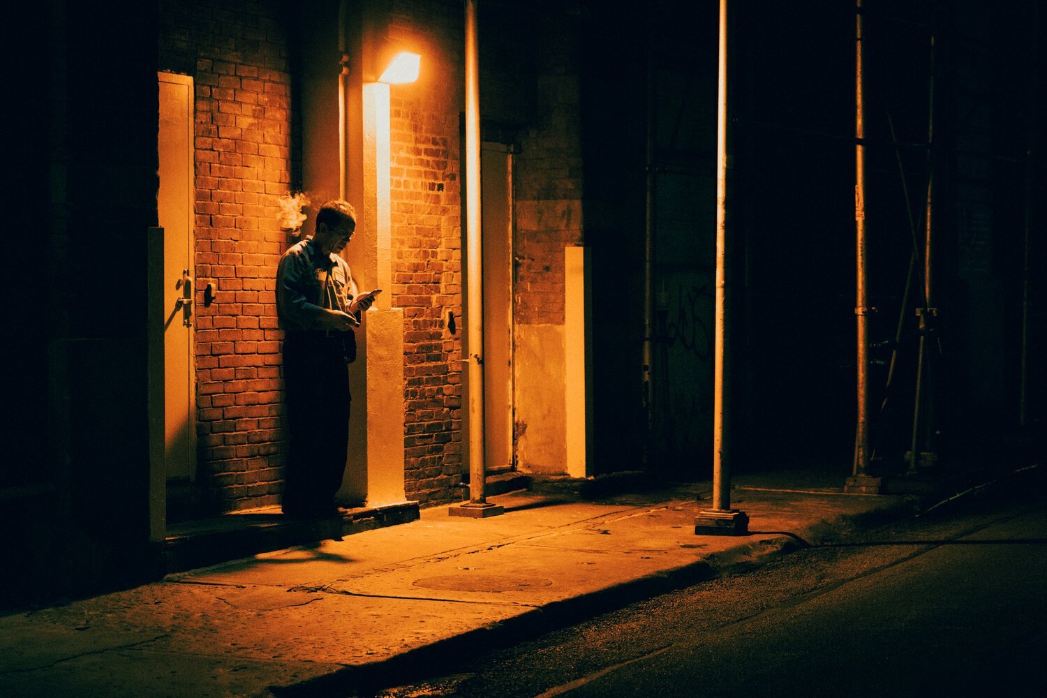 A person standing alone at night under a streetlight, leaning against a brick wall and looking at their phone.