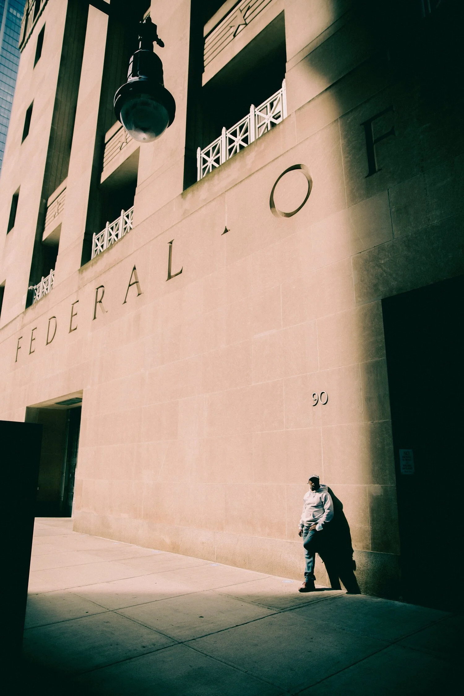 A man wearing a white hoodie and face mask leaning against a beige stone wall in front of the Federal building, with large cut-out letters on the wall reading 'FEDERAL'.