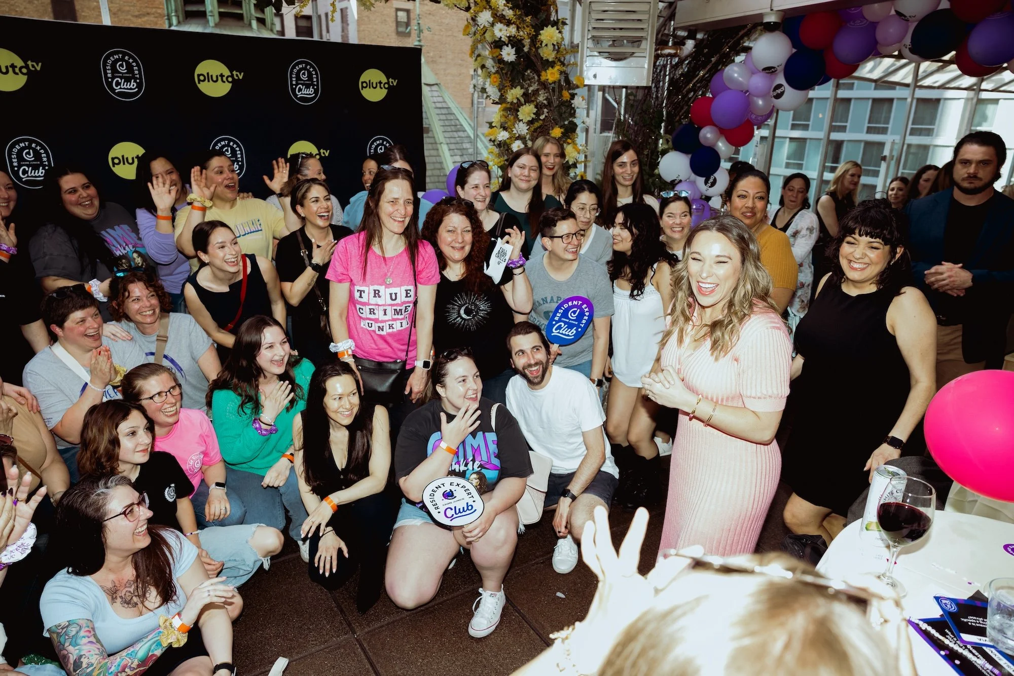 A large group of women gathered together at an indoor event decorated with balloons and flowers, smiling, laughing, and holding signs, with some wearing colorful clothing and accessories.