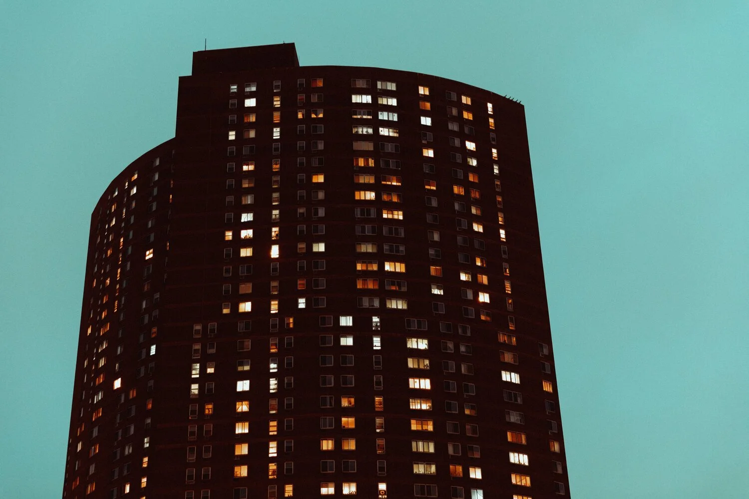 A tall, dark apartment building with lit windows against a teal evening sky.
