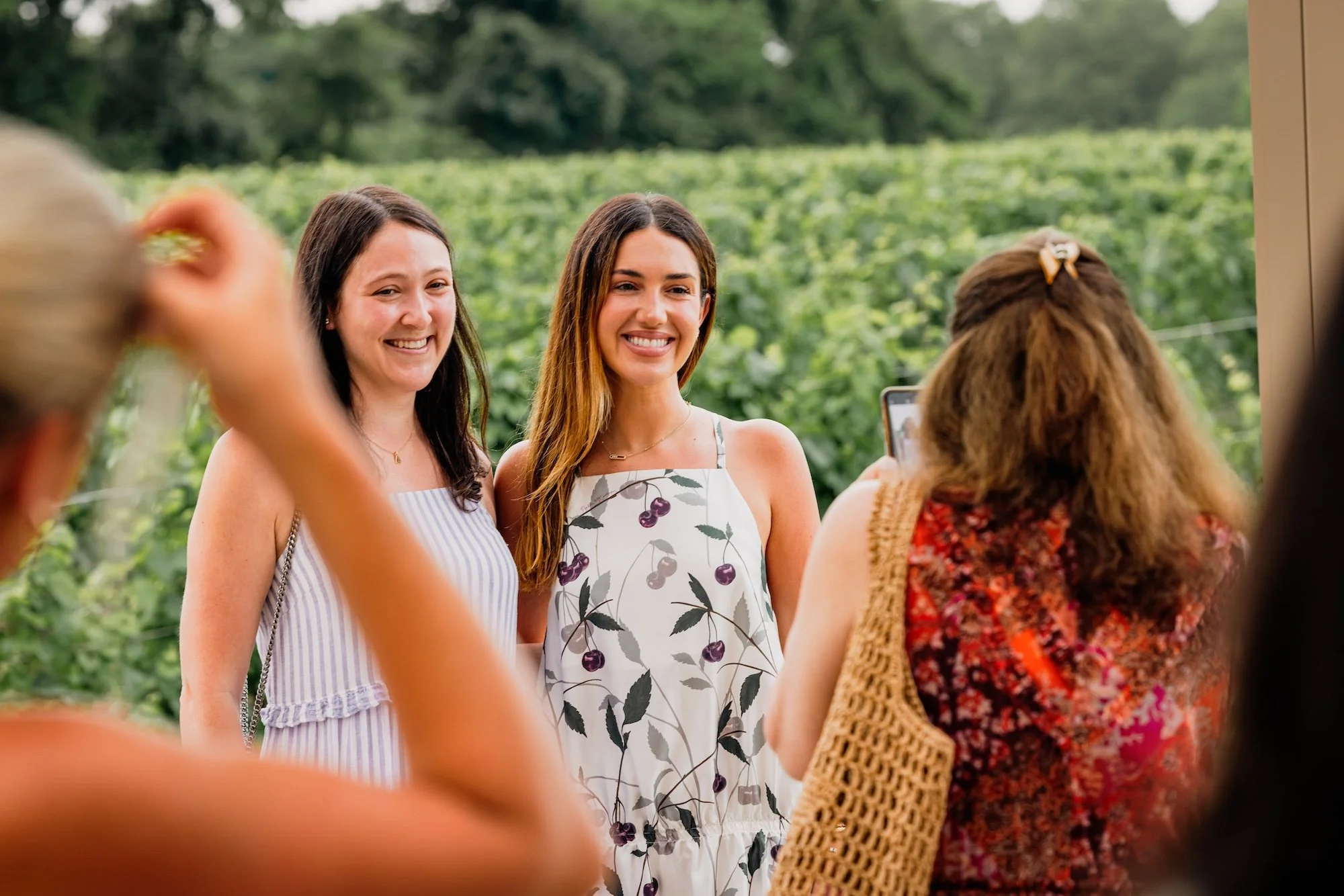 Two women standing outdoors in front of a green landscape, smiling for a photo taken by a person with long, curly hair wearing a patterned top.