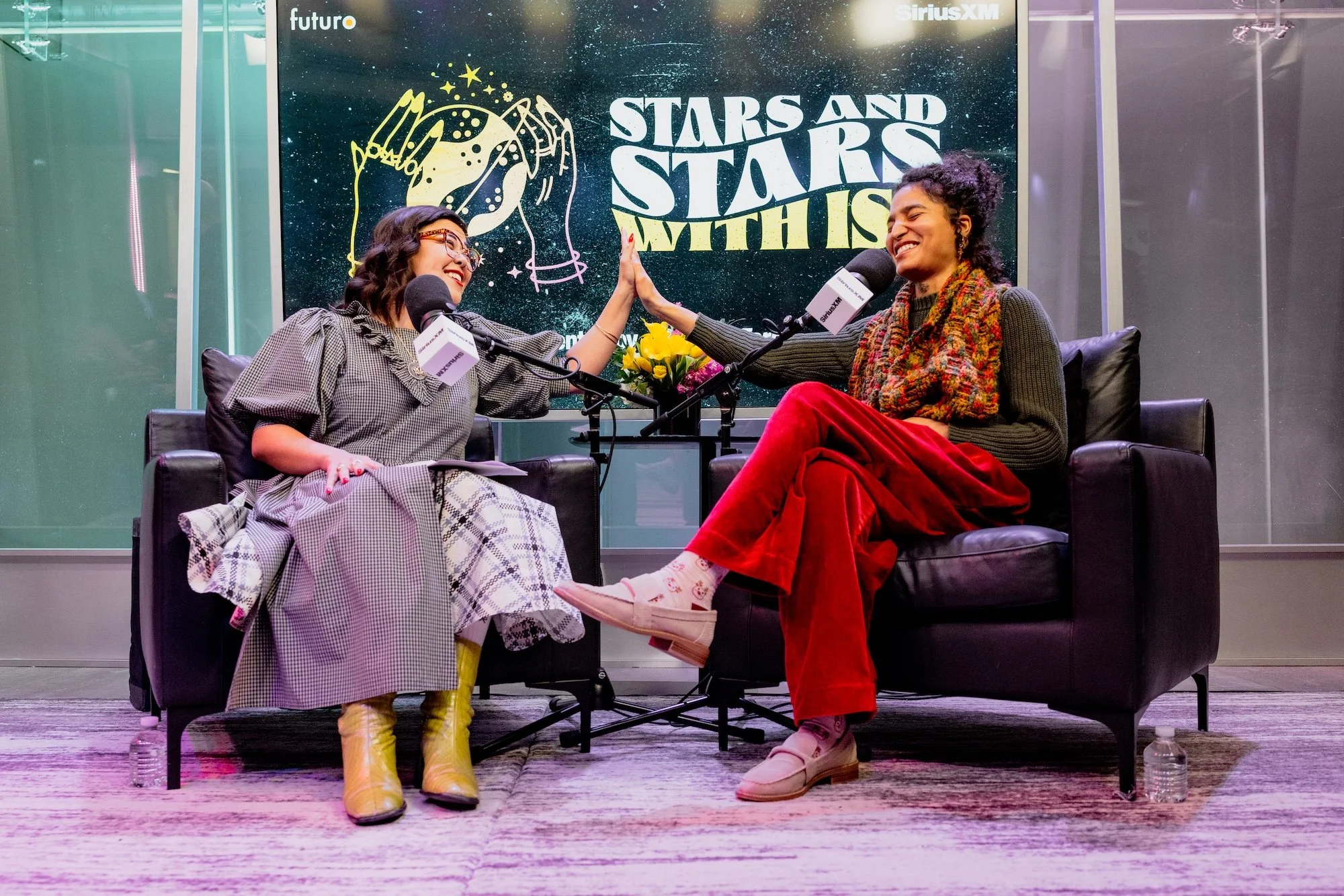 Two women seated on black armchairs on a stage, engaged in a conversation, high-fiving each other, with a backdrop displaying the text "Stars and Stars with" and a starry galaxy illustration, flowers on a table between them, microphones, and water bo