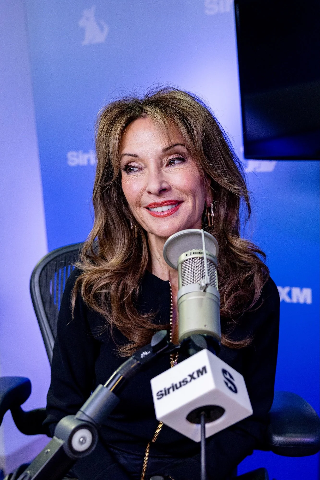 A woman with long, wavy brown hair smiling during a radio interview at SiriusXM studio, with a microphone in front of her and SiriusXM and TV screens in the background.