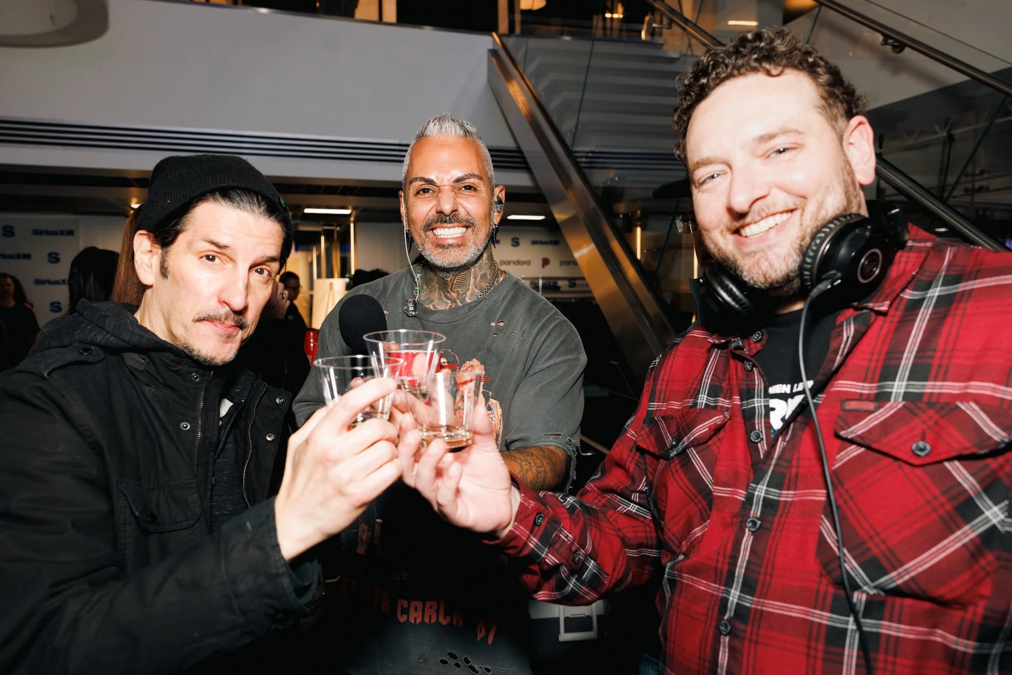 Three men are in a social setting, smiling and clinking clear shot glasses. The man on the left has long hair, a mustache, and wears a black beanie and jacket. The man in the middle has gray hair, tattoos on his neck and arms, and is wearing a gray d