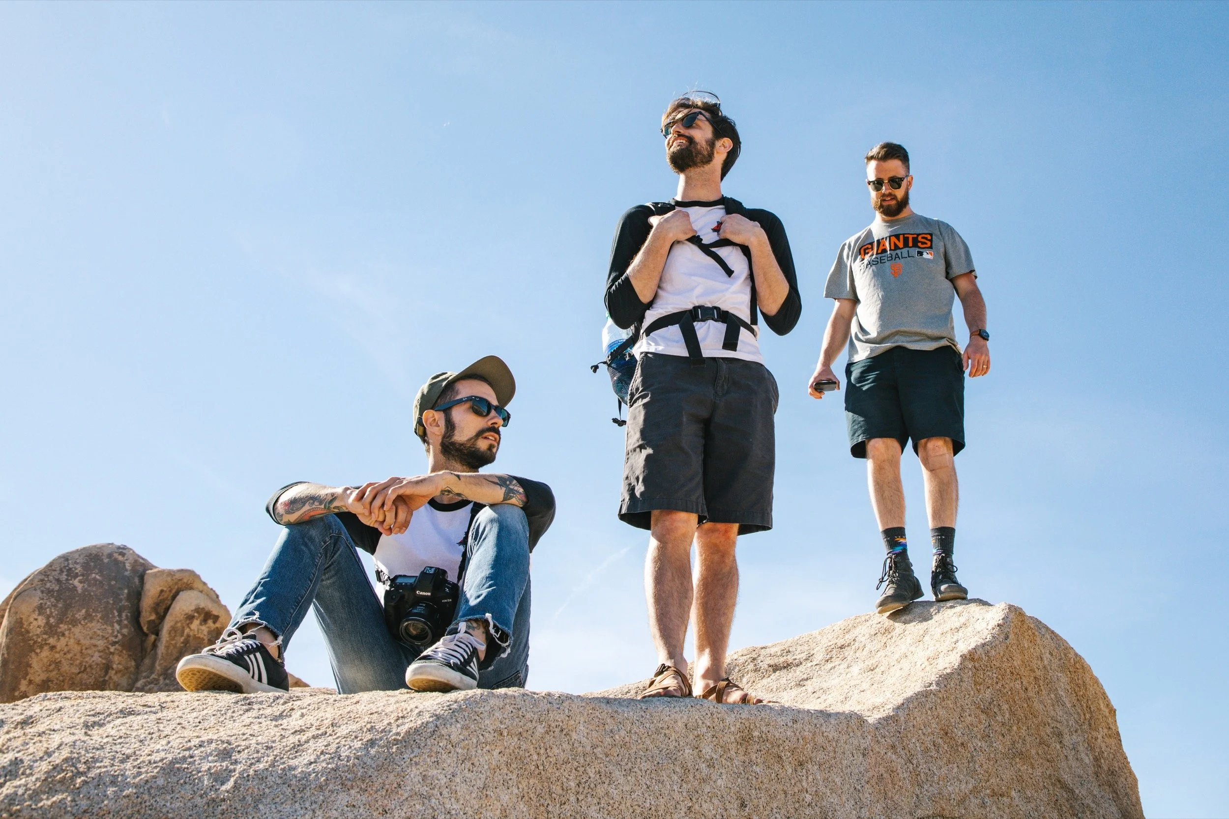 Three men outdoors on a large rock under a clear sky, one sitting with a camera, two standing, all wearing casual clothing and sunglasses.