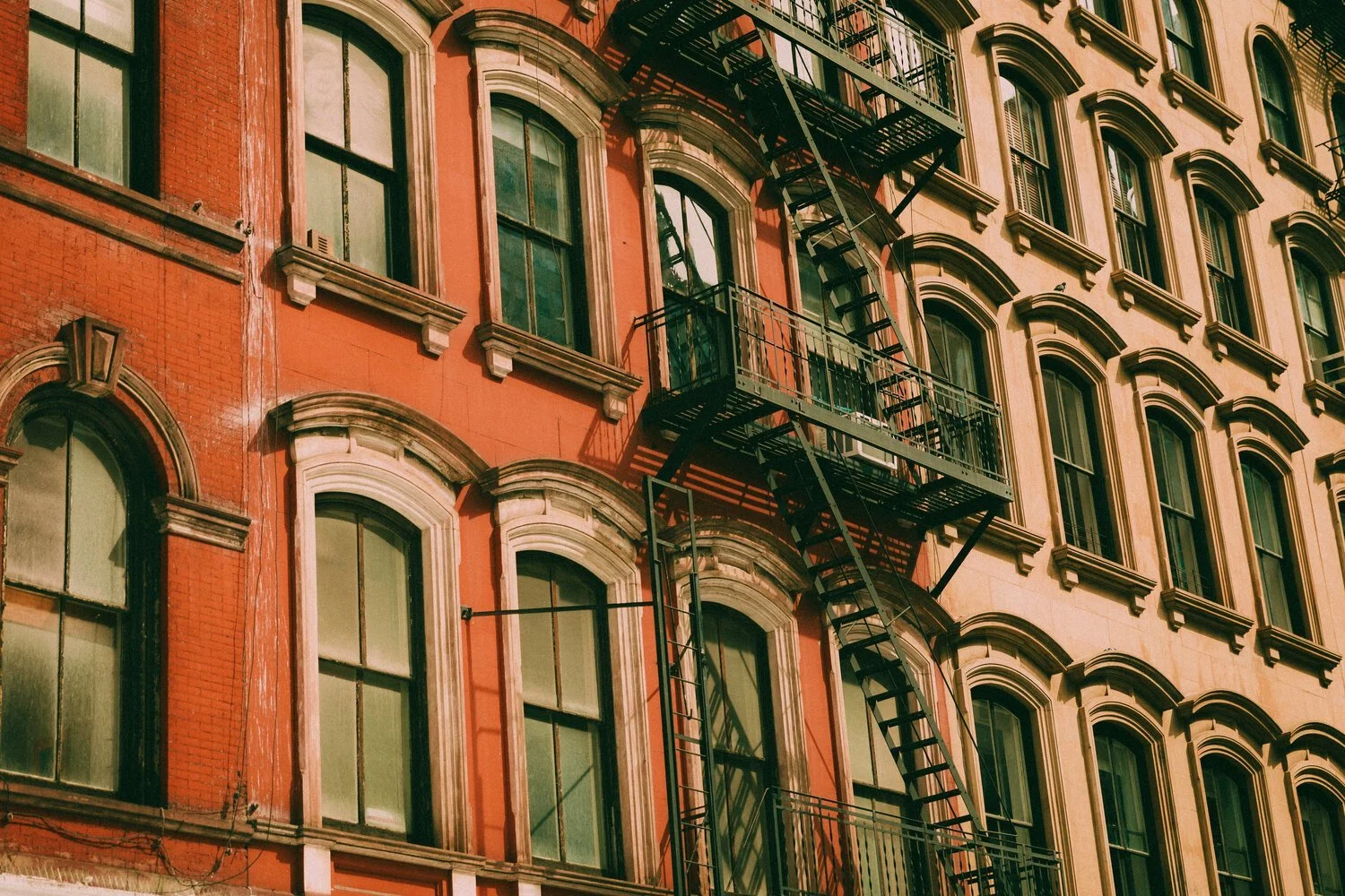 The image shows a multi-story building with large arched windows and a decorative brick facade. There is a black metal fire escape stairway attached to the exterior, extending from the lower to the upper levels, casting shadows on the building wall.