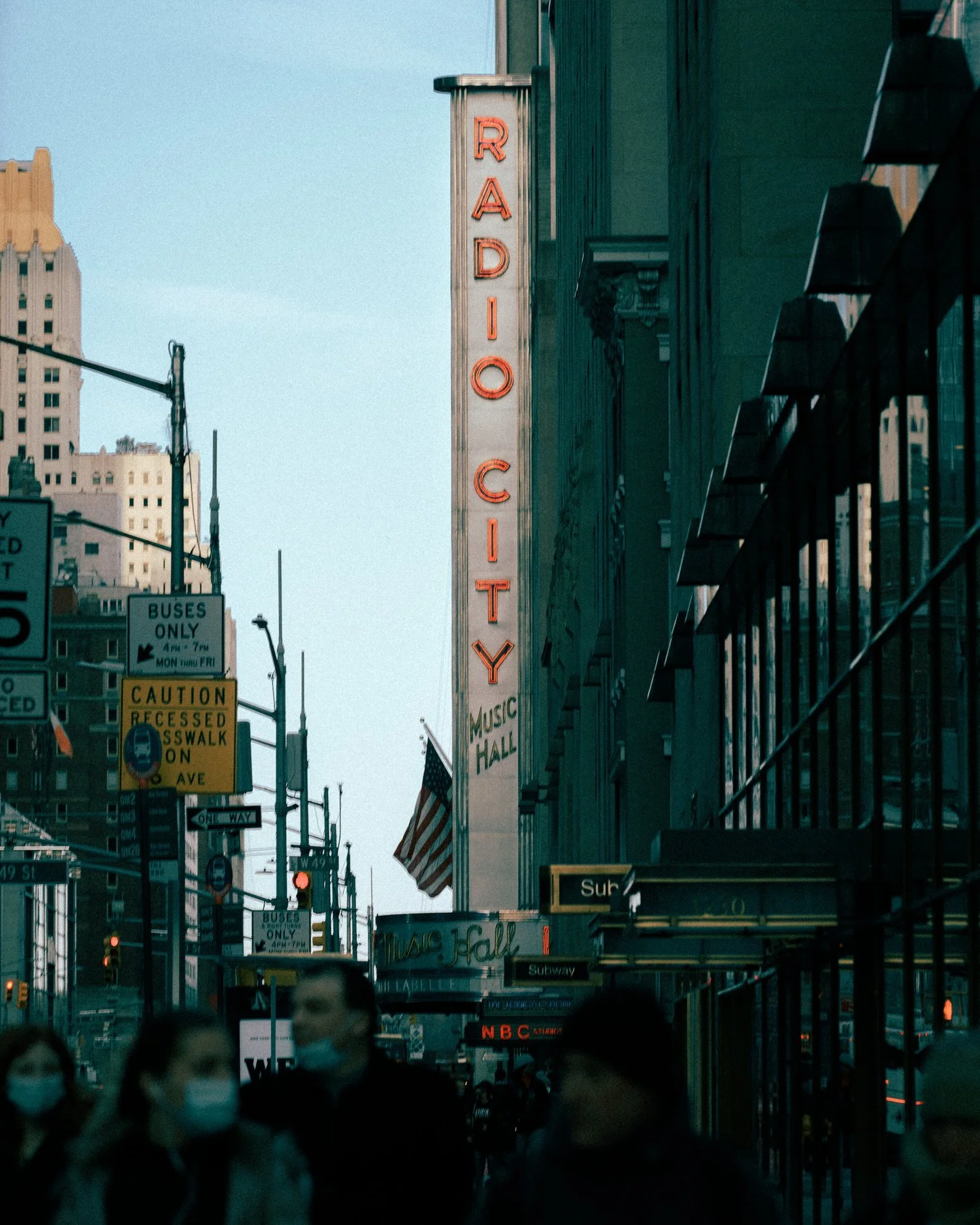 Street scene in New York City with illuminated 'Radio City' sign and people walking, some wearing masks, buildings, and signs for buses, subway, and music hall.