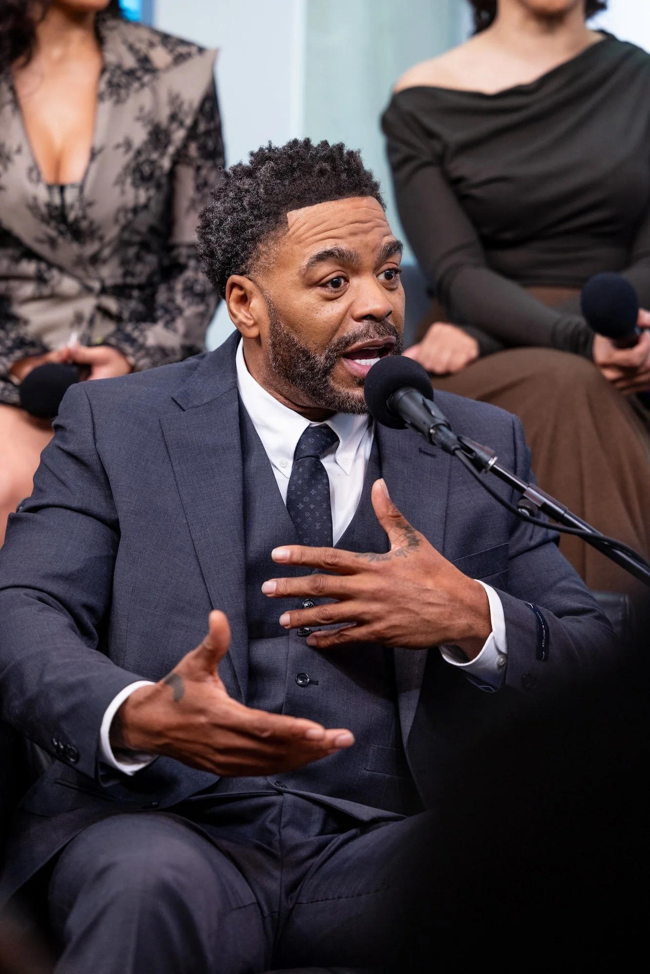 Man speaking into a microphone at a panel discussion, gesturing with his hand, with two women sitting behind him.